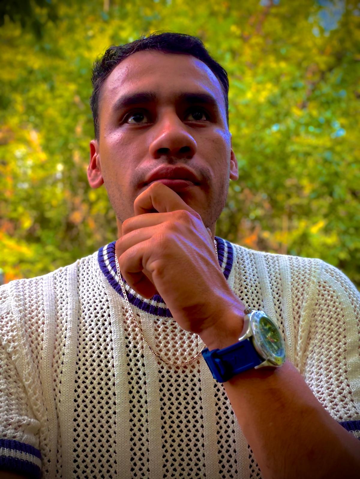 Man with short dark hair, wearing a white knit sweater with blue accents, resting his chin on his hand outdoors.