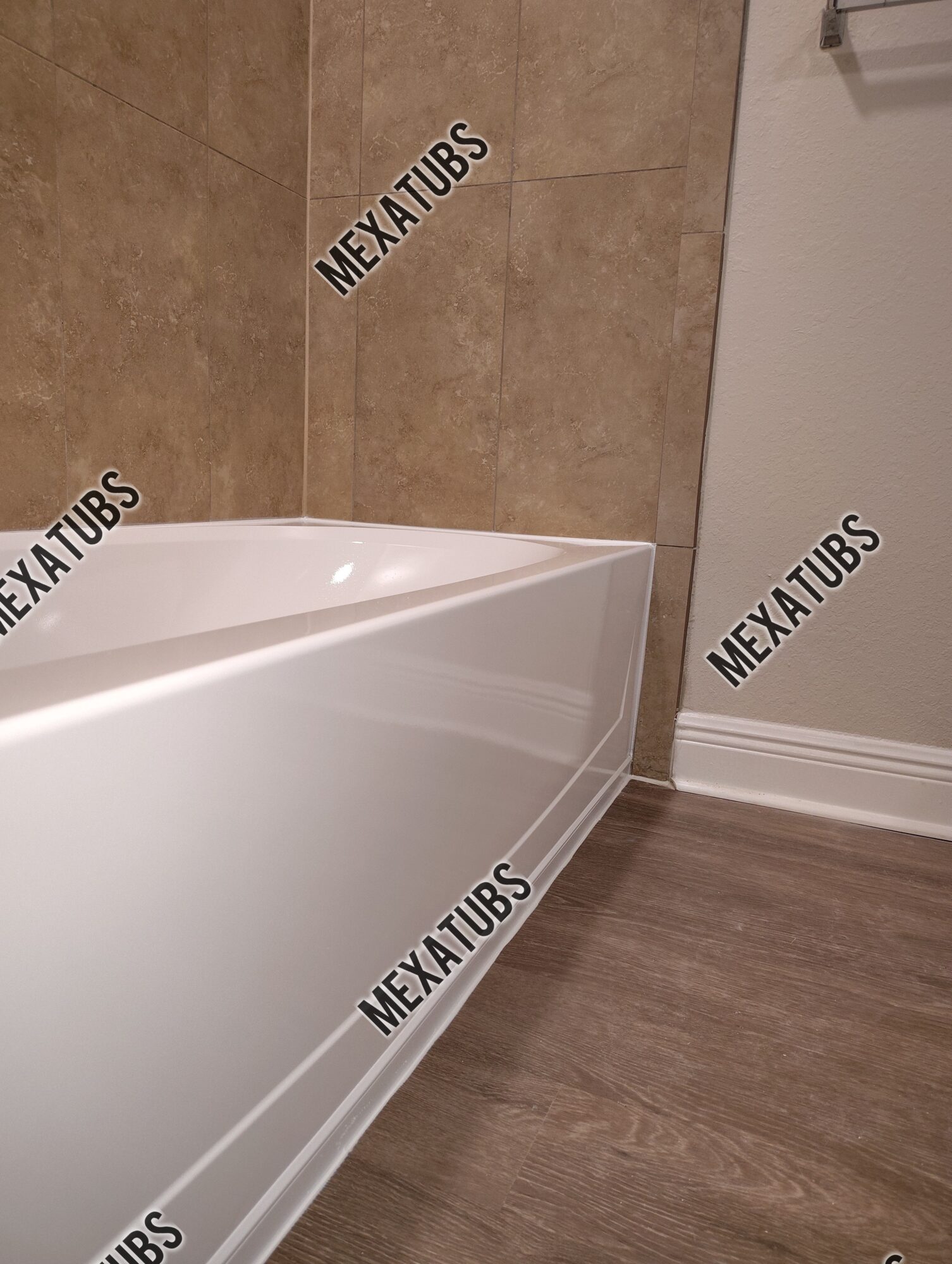 Corner of a bathroom with a white bathtub, beige tiled wall, and wooden floor, with a white wall and baseboard.