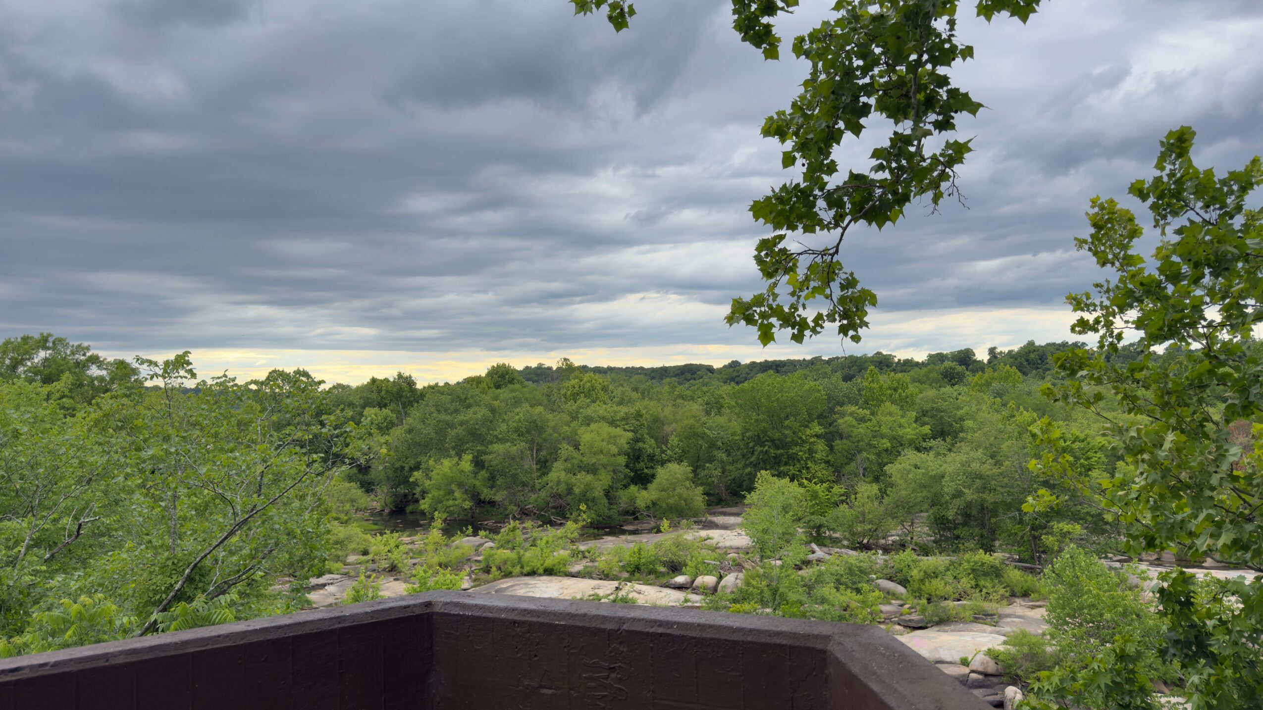 View of a cloudy sky over a green landscape with trees and a rooftop in the foreground.