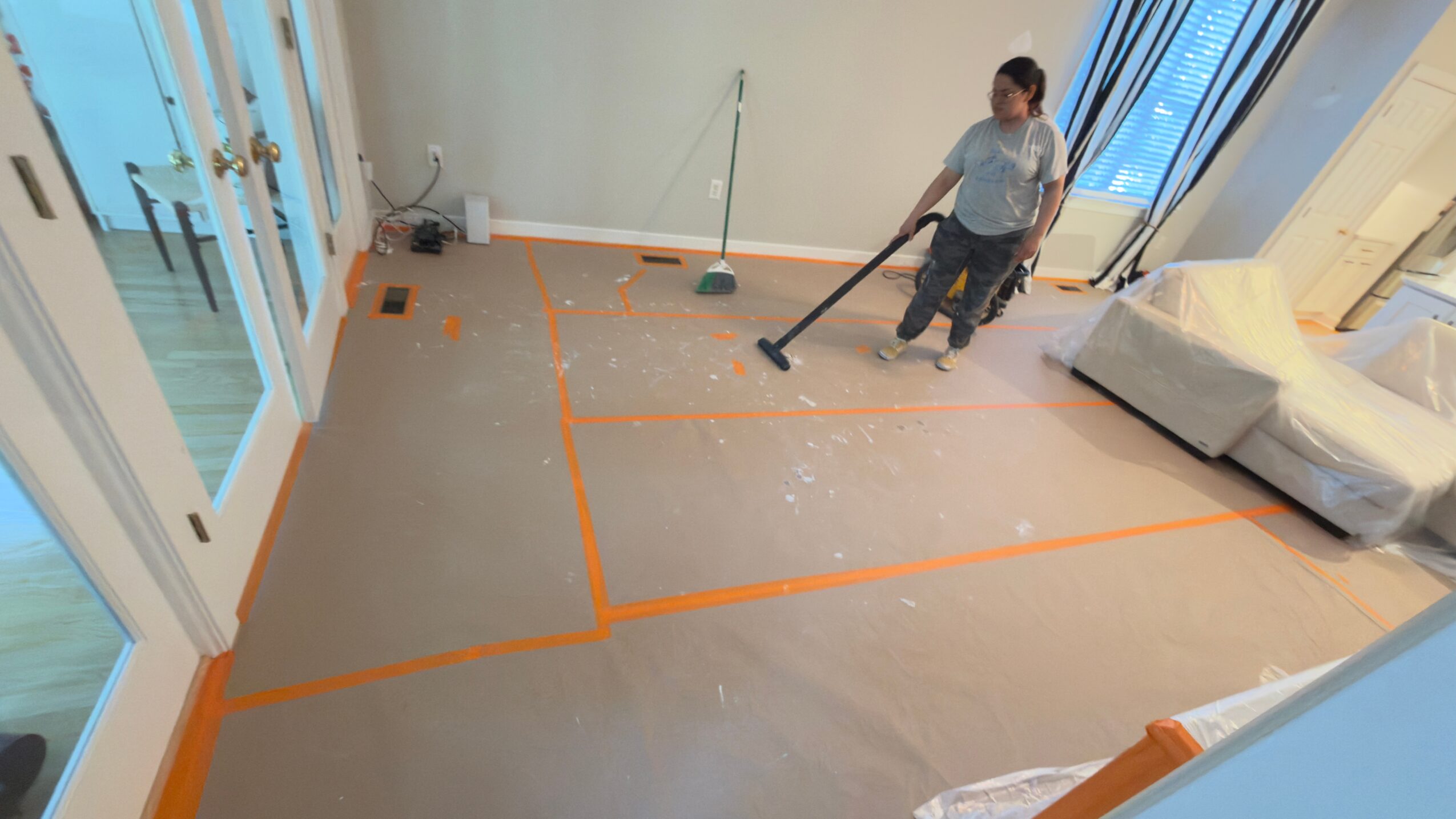 Person cleaning a room with orange tape on the floor, a mop, and a sofa, with large windows and a balcony door.