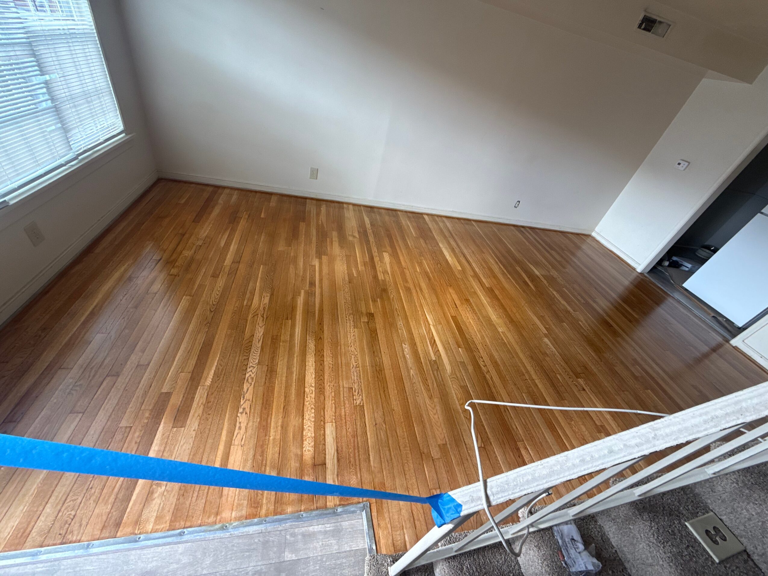 Empty room with wooden floor, white walls, and large window, viewed from a staircase landing.