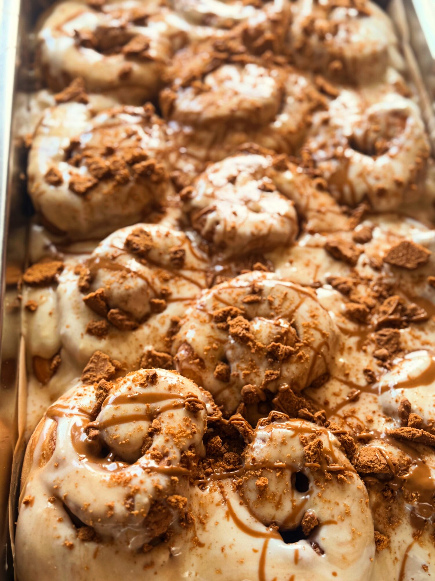 Close-up of cinnamon rolls topped with icing and crumbled cookies or nuts, arranged in a tray.