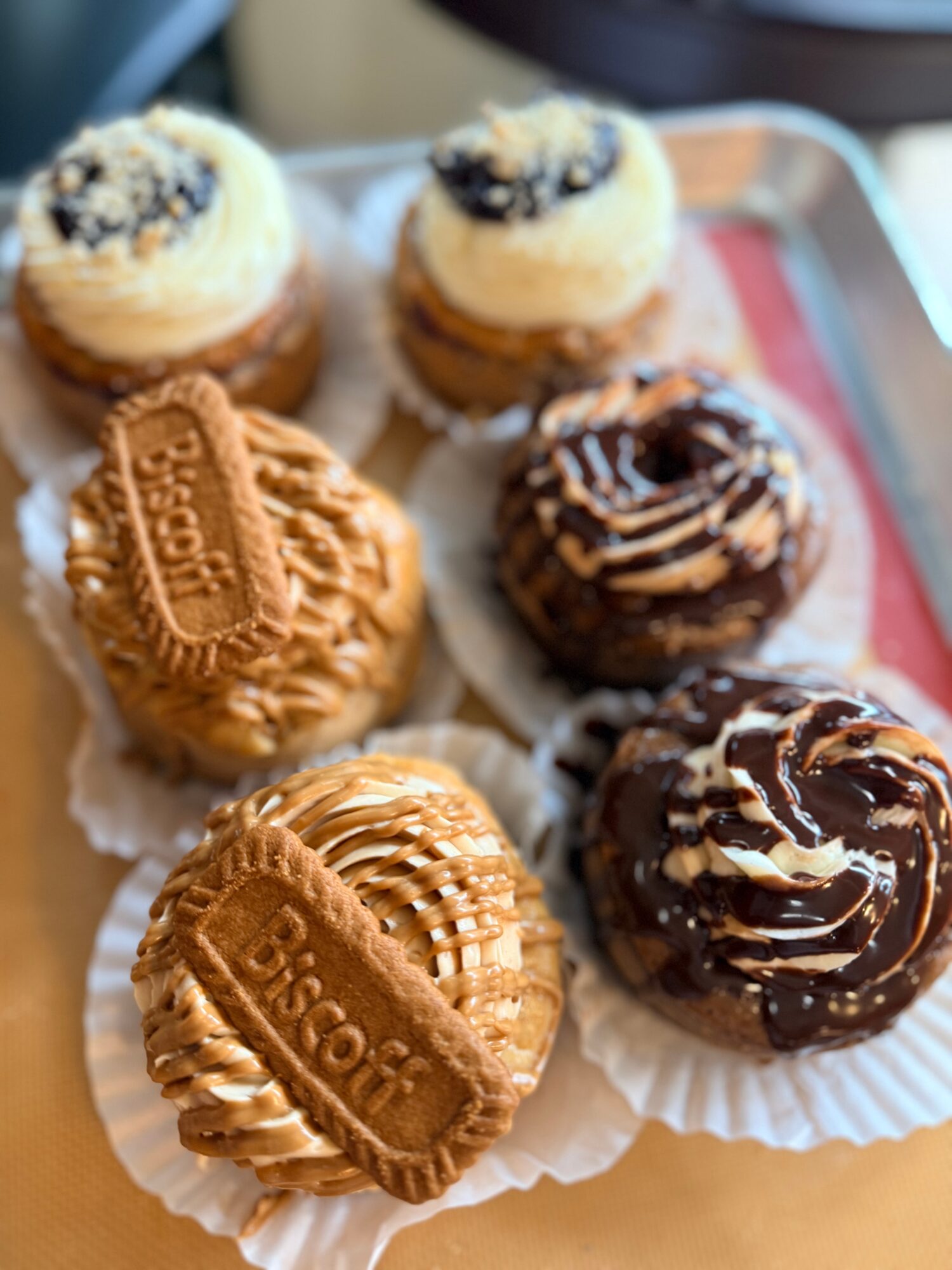 Six assorted cupcakes and cookies on a tray, some with chocolate and others with vanilla frosting, in paper cups.