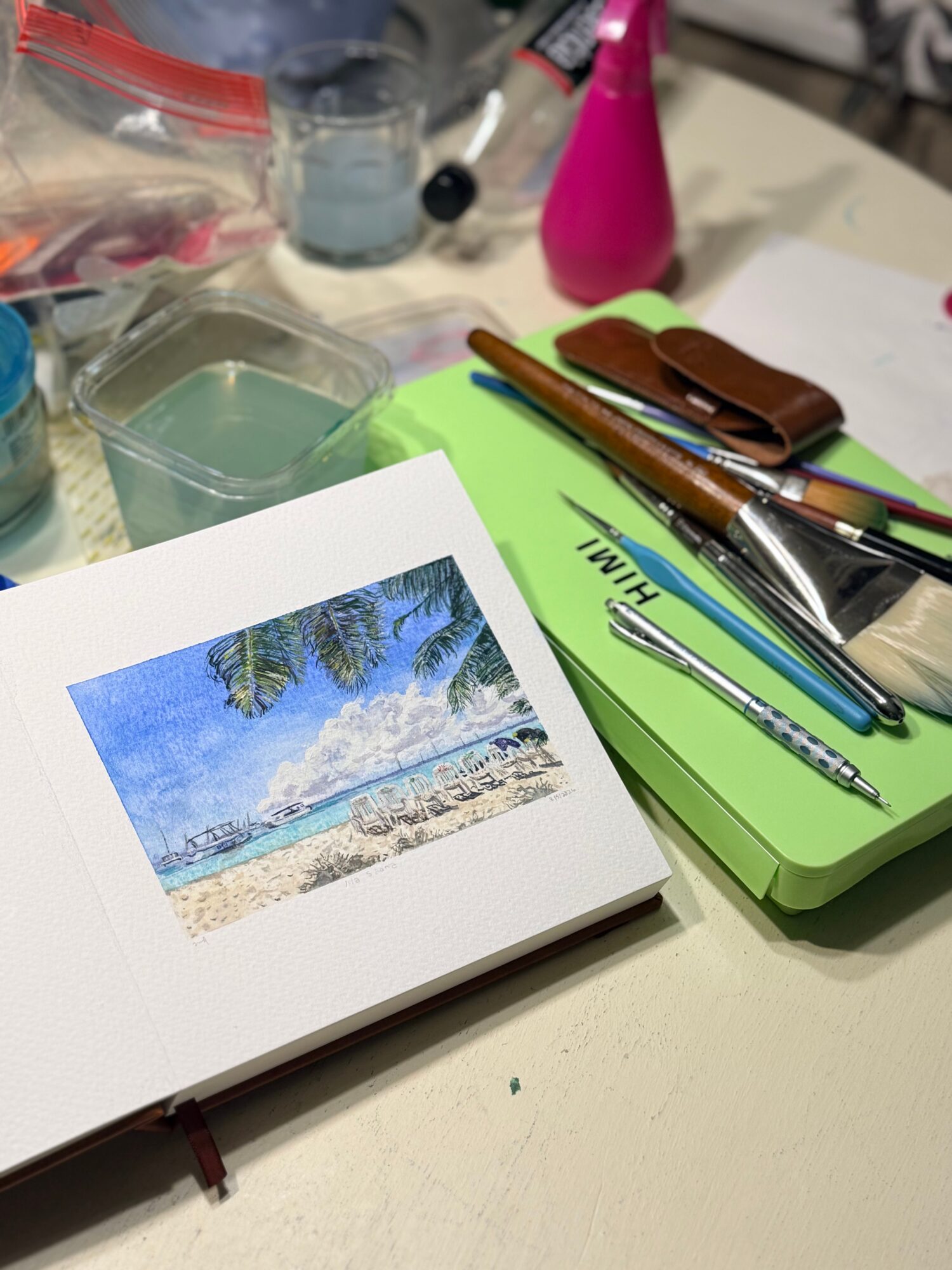 Photograph of a beach with blue sky, palm trees, and ocean, printed on a small card on a cluttered desk.