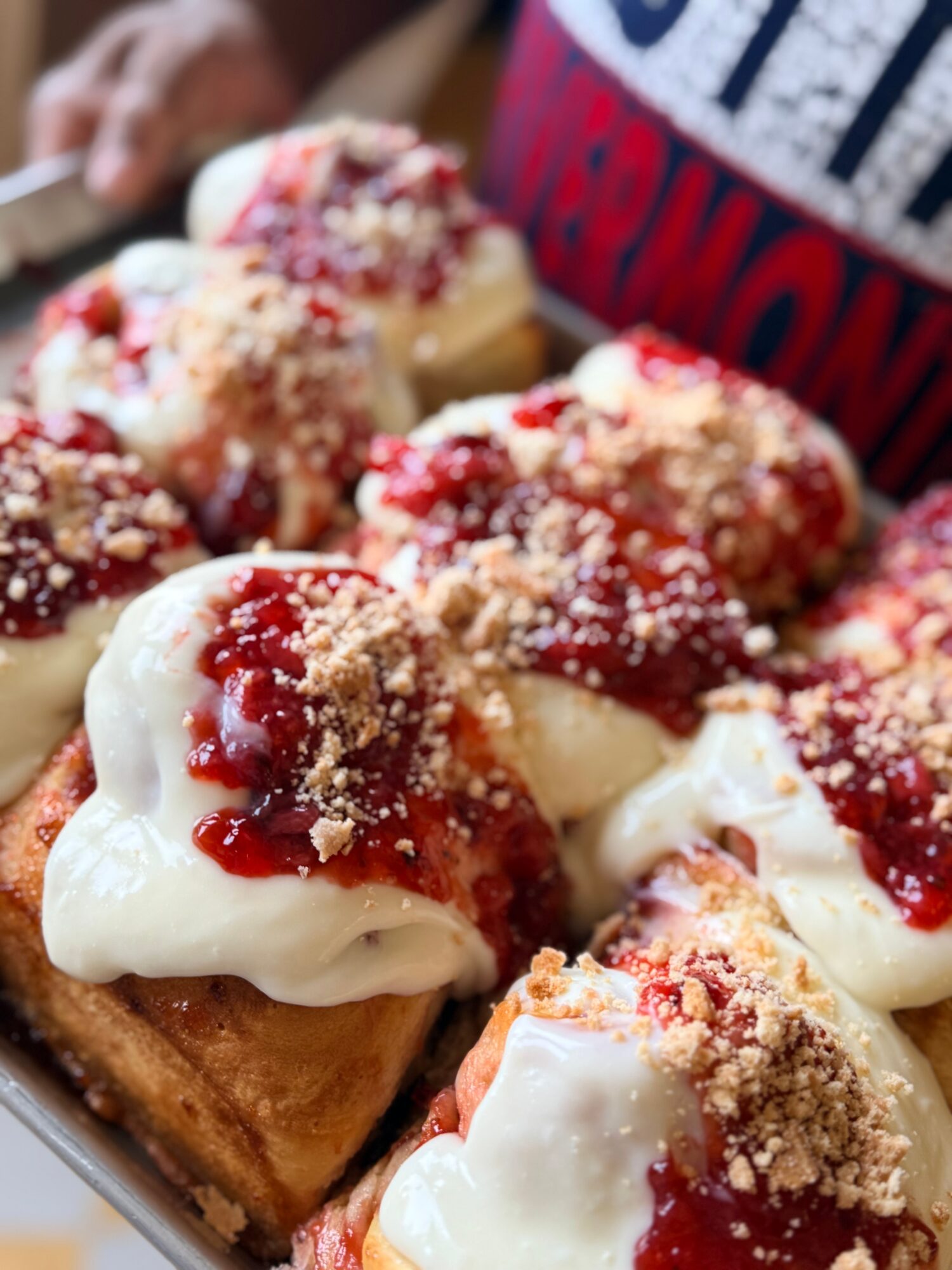 Close-up of dessert squares topped with whipped cream, red fruit sauce, and crumbly topping, with a person wearing a red and blue shirt in background.