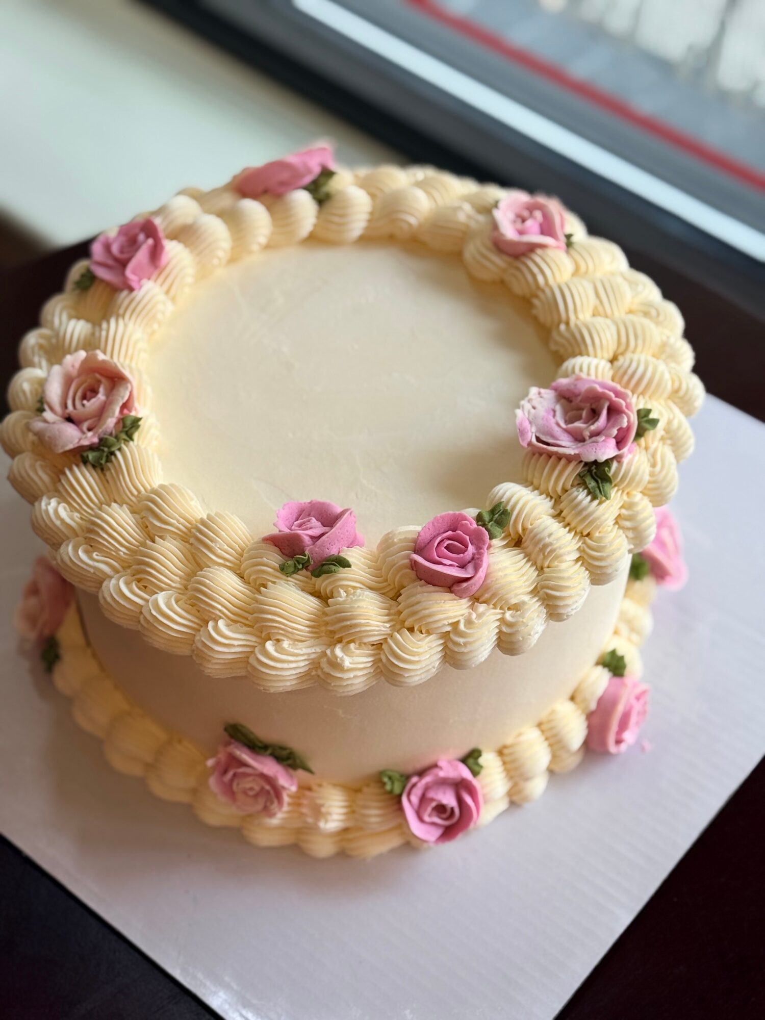 Round cake decorated with cream swirls and pink roses, placed on a white surface near a window.