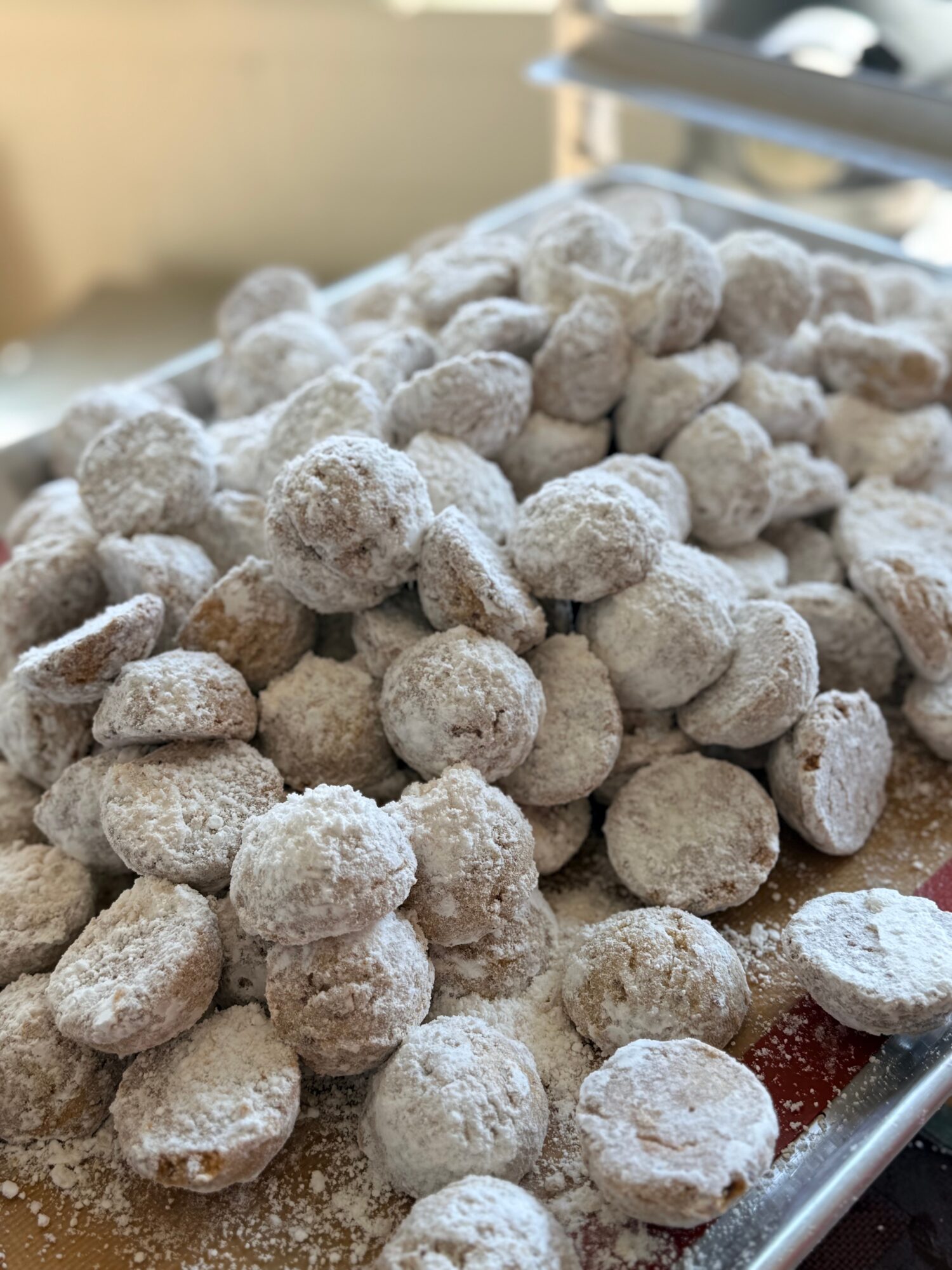 Pile of small, round, powdered sugar-coated cookies or confections on a tray, with a blurred background.
