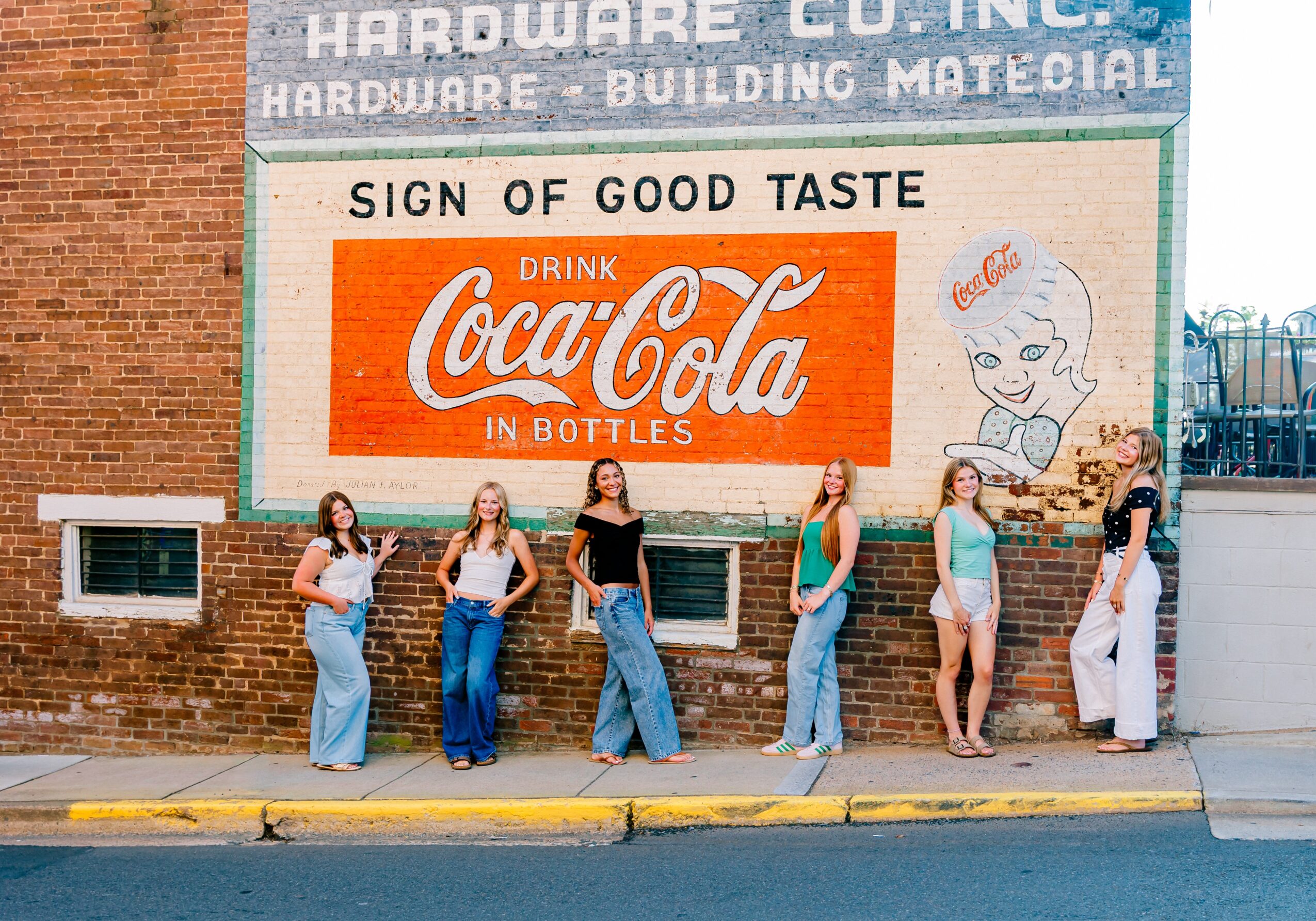 Six women stand in a line on the sidewalk in front of a brick wall with vintage advertisements, facing forward.