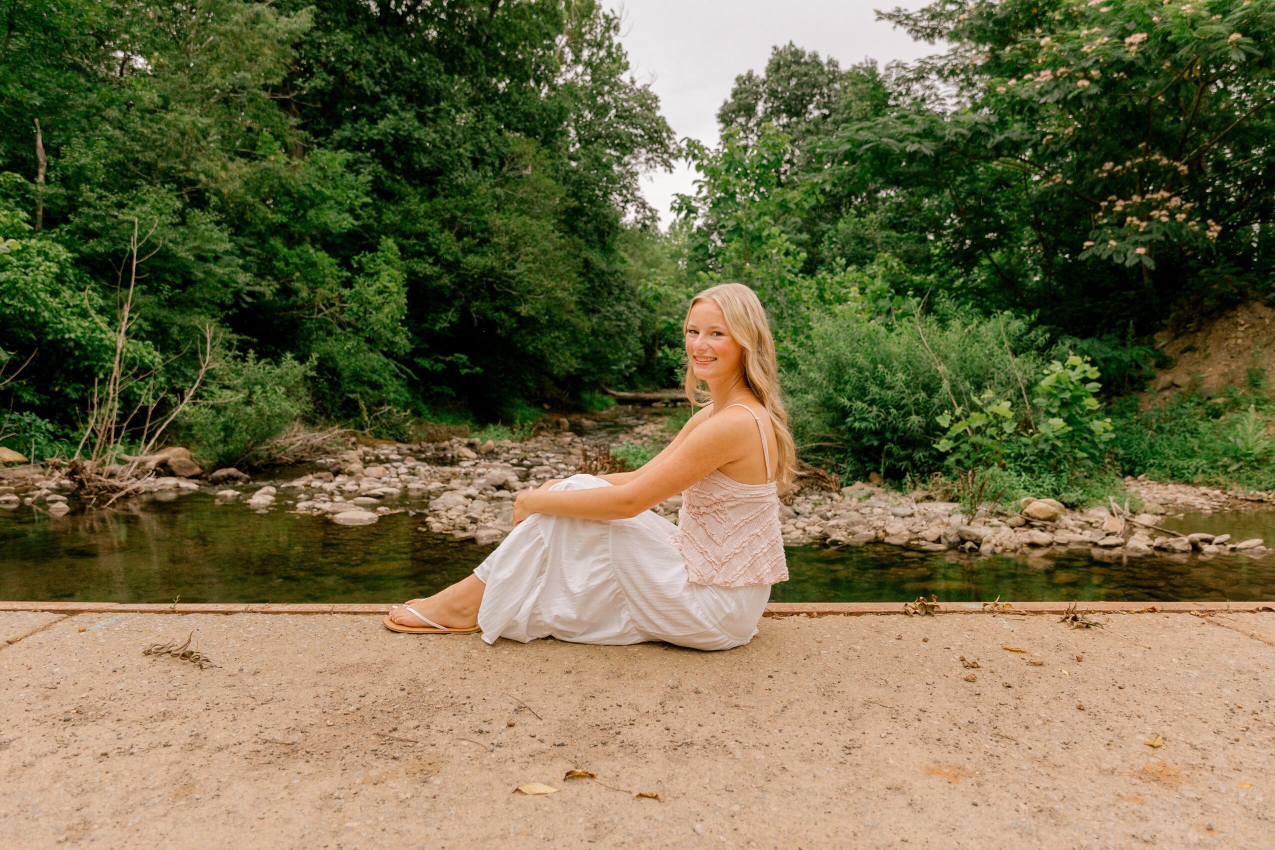 Woman sitting on a sandy riverbank with trees and water in the background, smiling and looking to the side.