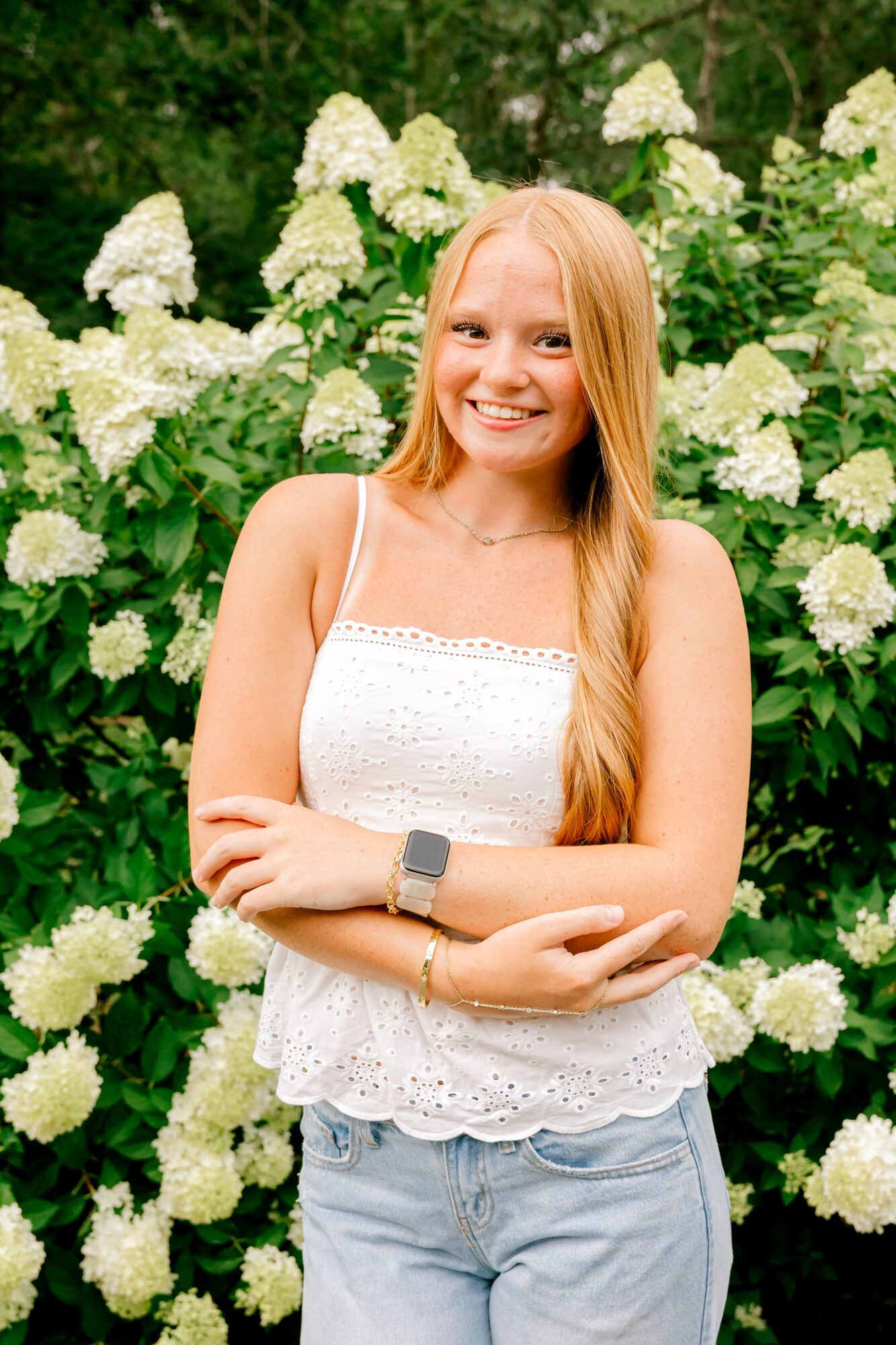 Young woman with long blonde hair smiling outdoors, standing in front of white hydrangea flowers, wearing a white sleeveless top and jeans.
