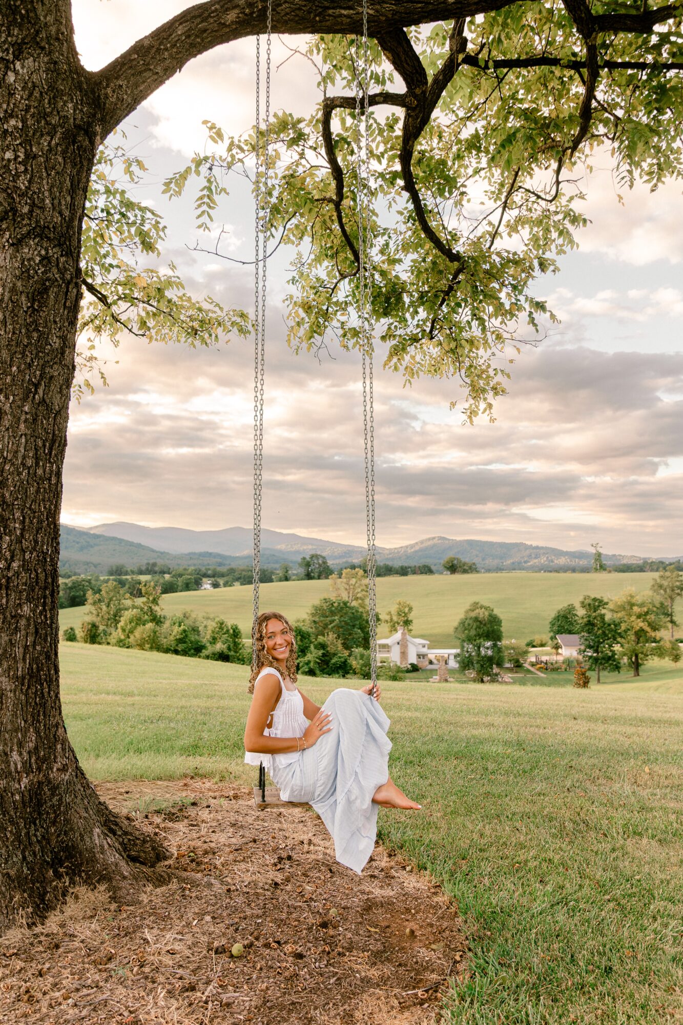 Woman sitting on a swing hanging from a large tree in a grassy field with a cloudy sky and distant hills.