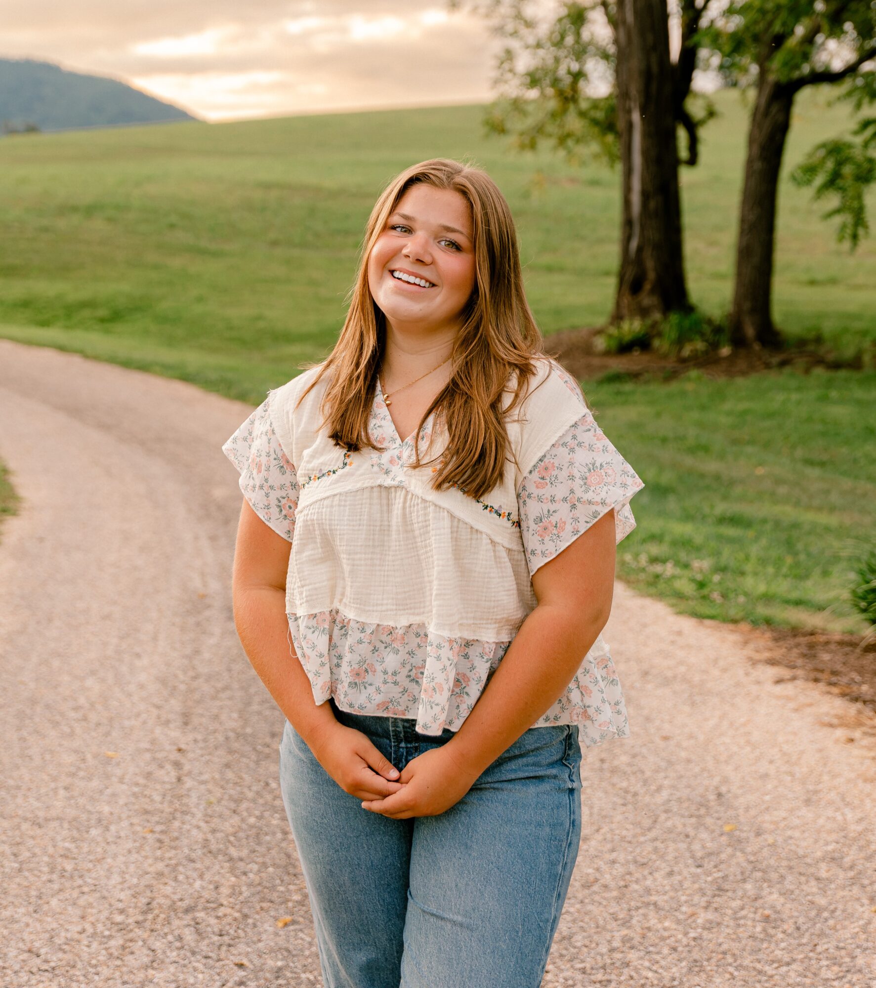 Smiling young woman standing outdoors on a dirt path with trees and grassy hills in background.