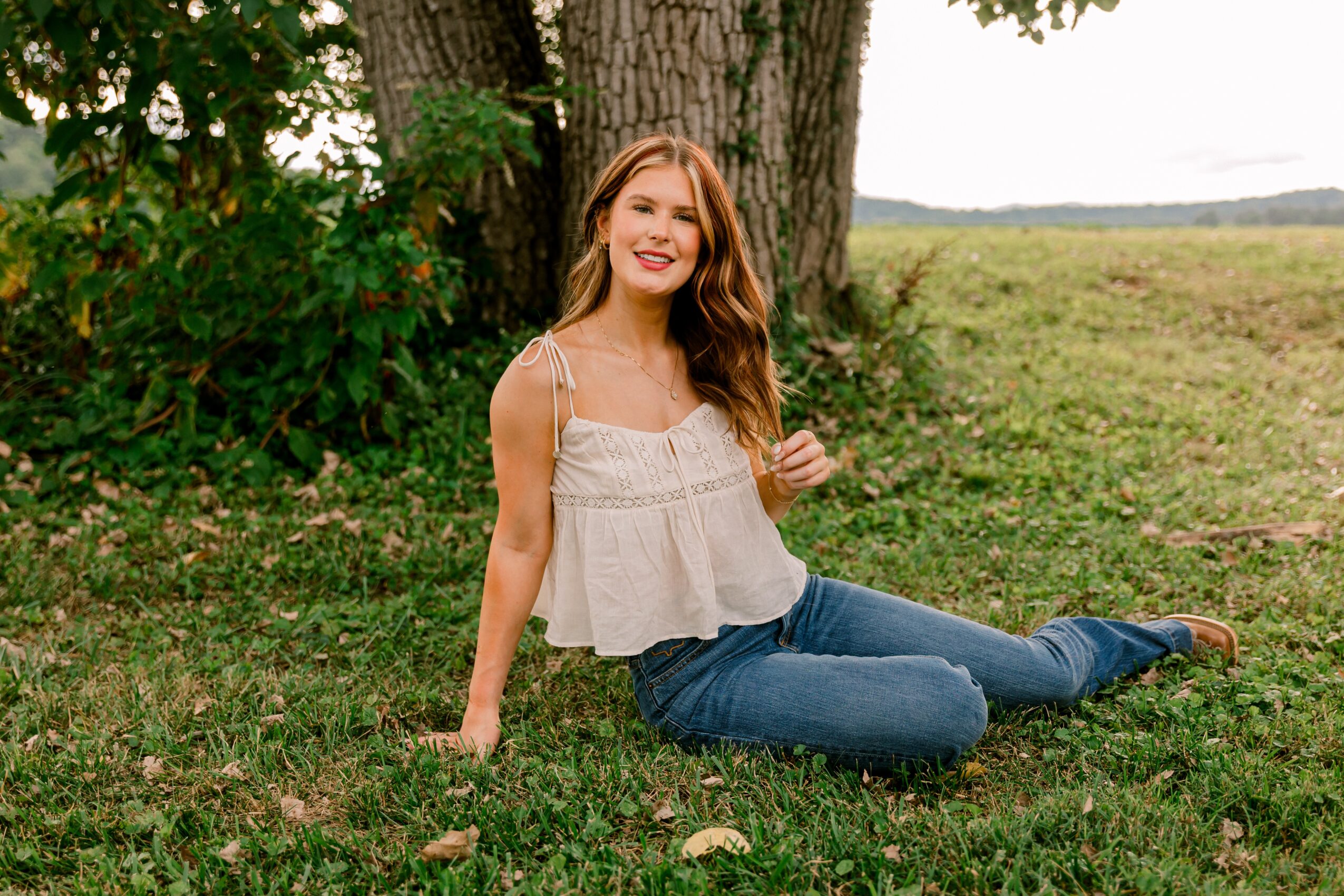 Young woman with long hair sitting on grass near a tree, smiling outdoors, wearing a white top and jeans.