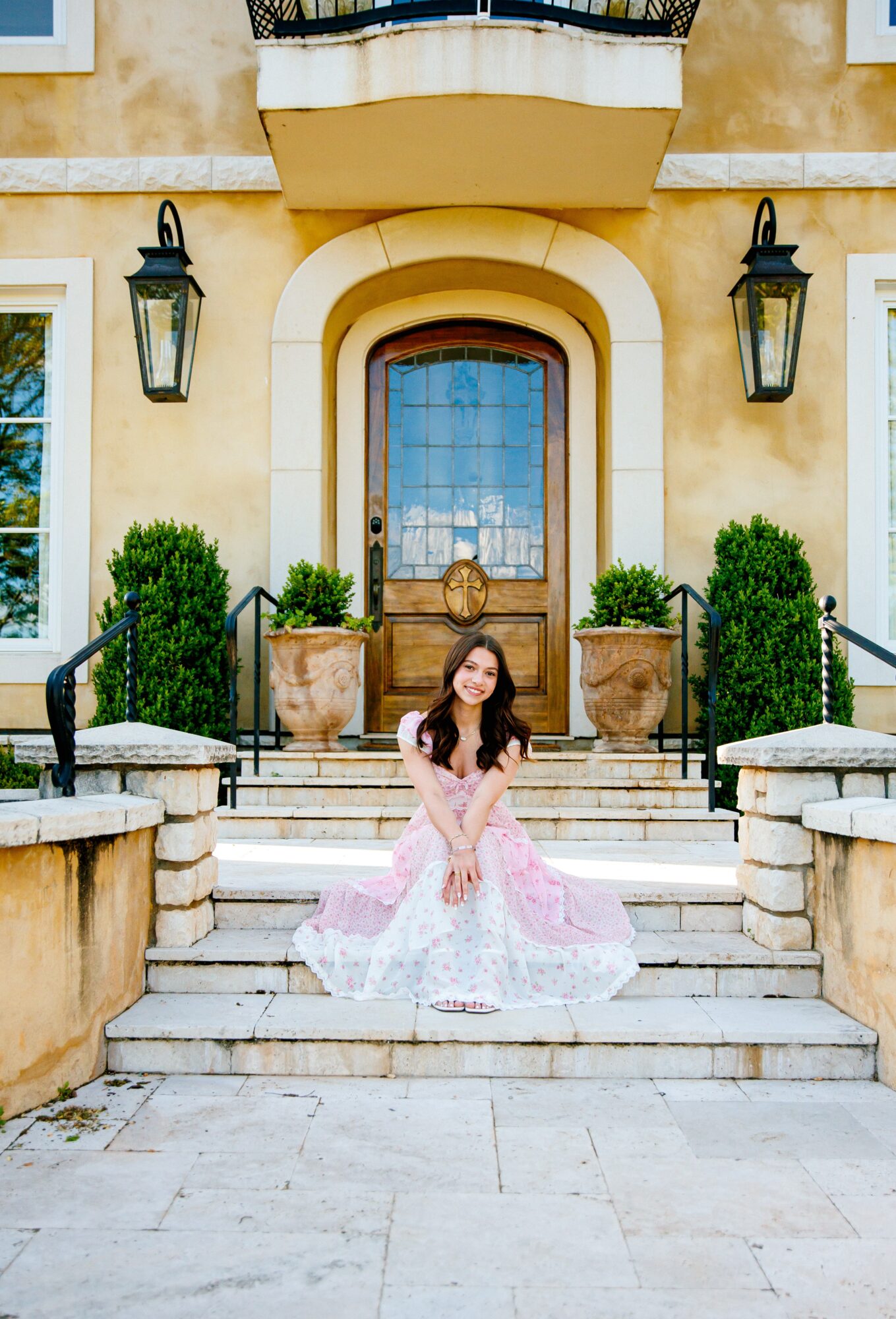 Young woman in a pink dress sitting on steps in front of a yellow building with potted plants and lanterns.