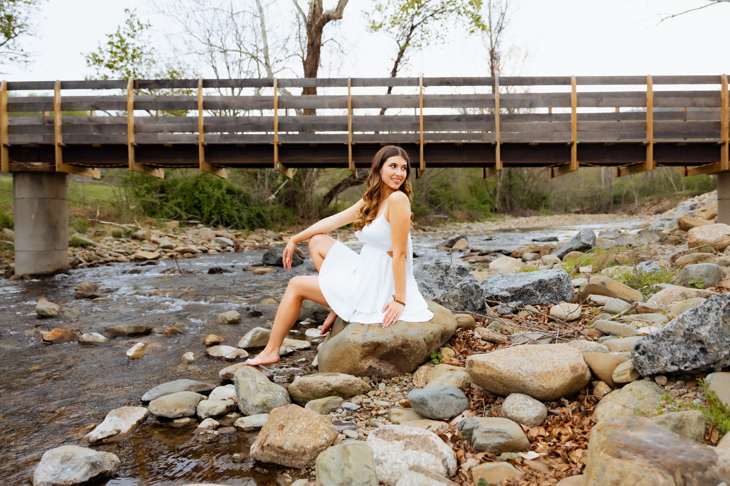 Woman in white dress sitting on rocks in a creek under a bridge, trees in background.