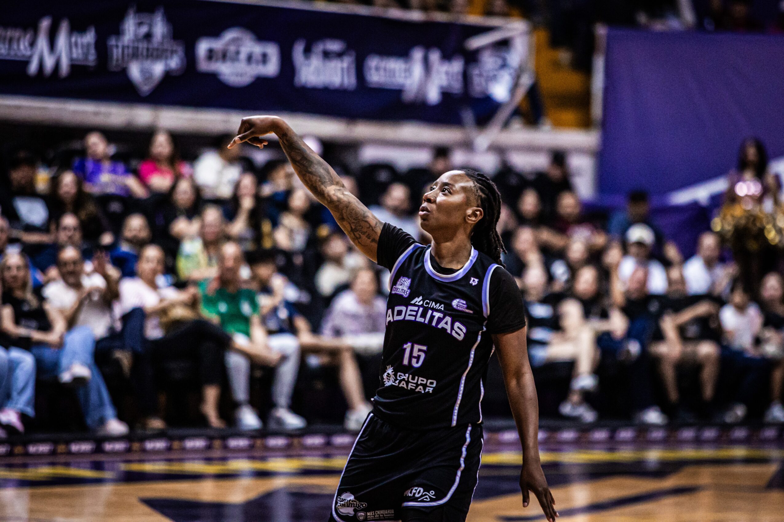 Basketball player in black uniform with number 15 prepares to shoot, audience seated in background.