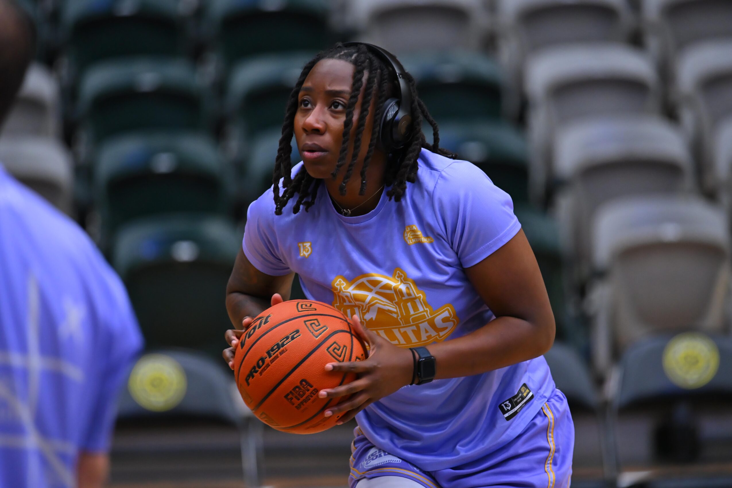 Young woman with dreadlocks holding a basketball on a sports court, wearing a purple sports uniform, with empty stadium seats in background.