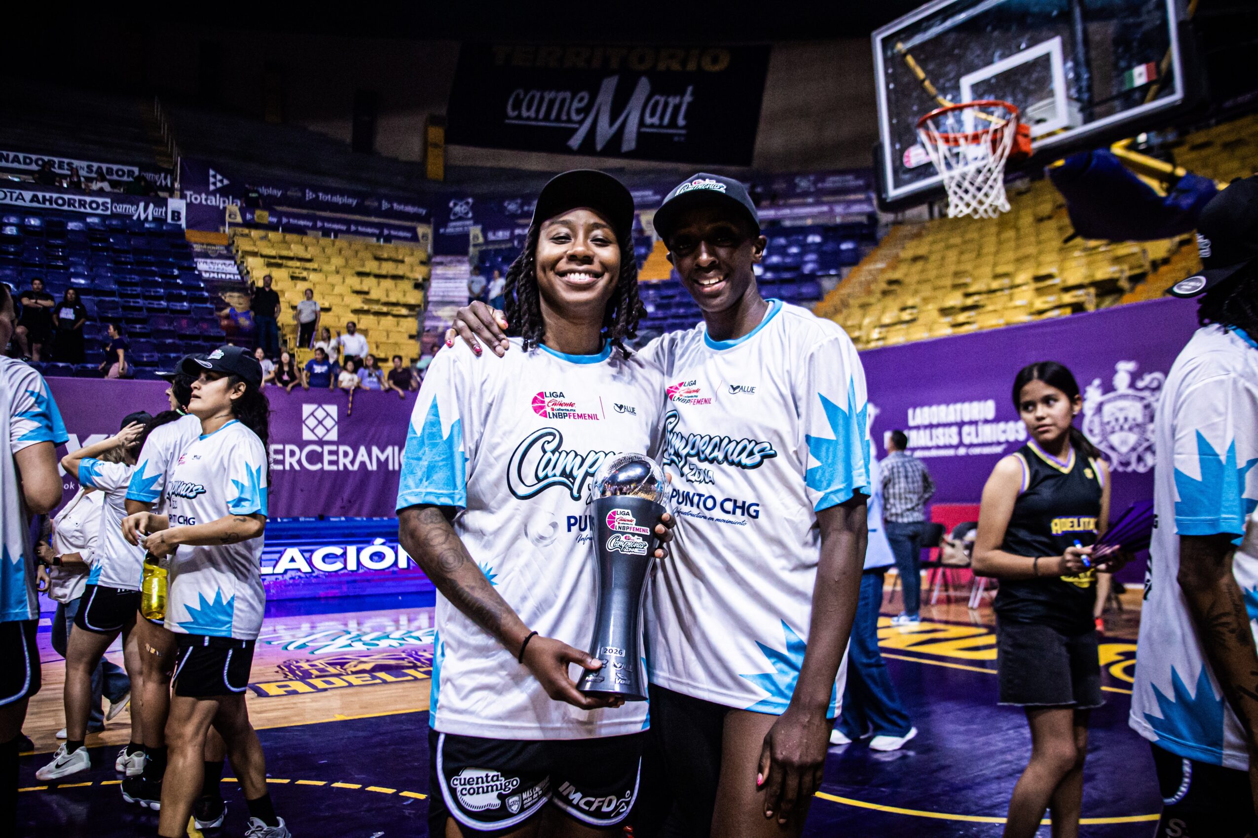 Two smiling women in basketball jerseys stand together on a court, holding a trophy, with other players and spectators in the background.