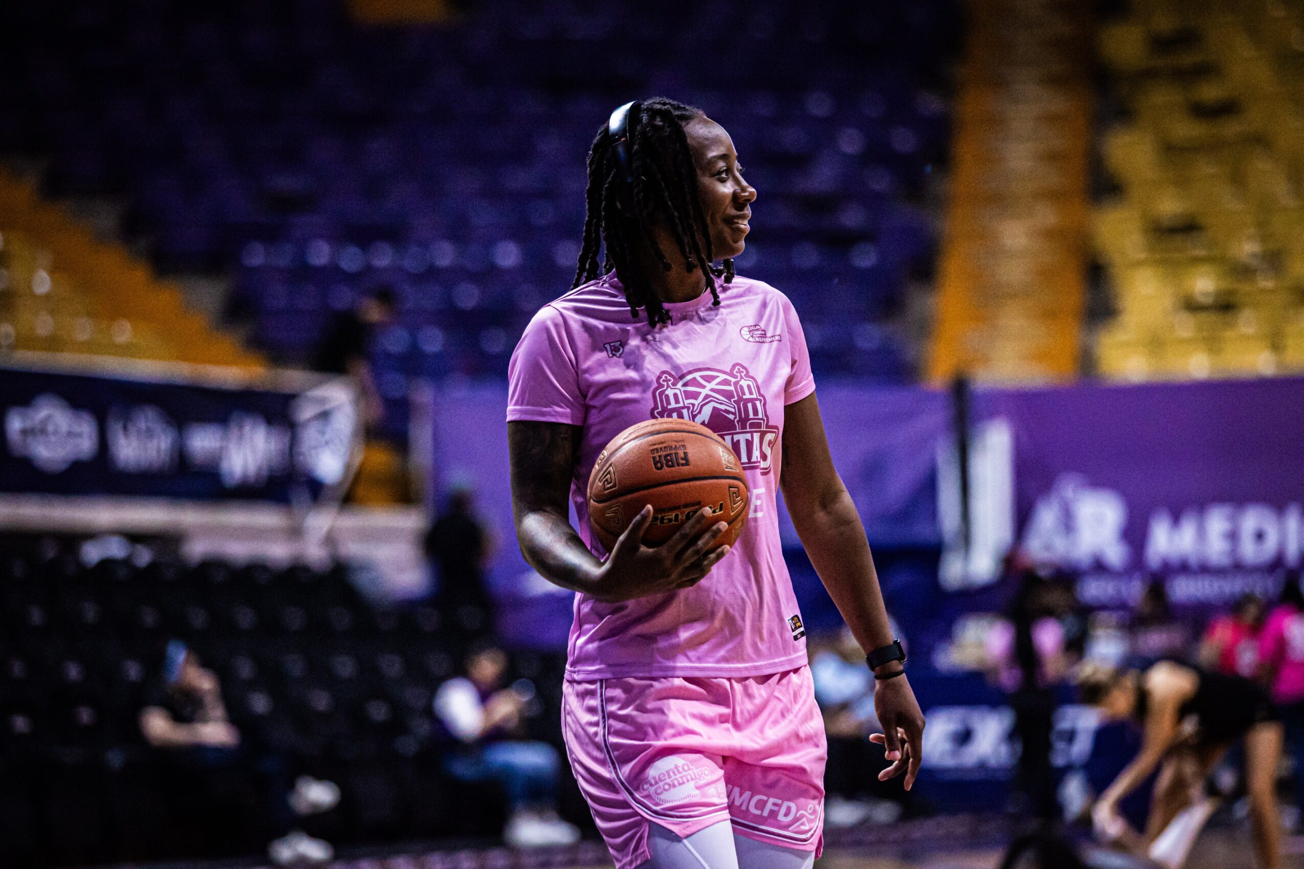 Woman in pink sportswear holding a basketball on a basketball court, with empty seats in the background.