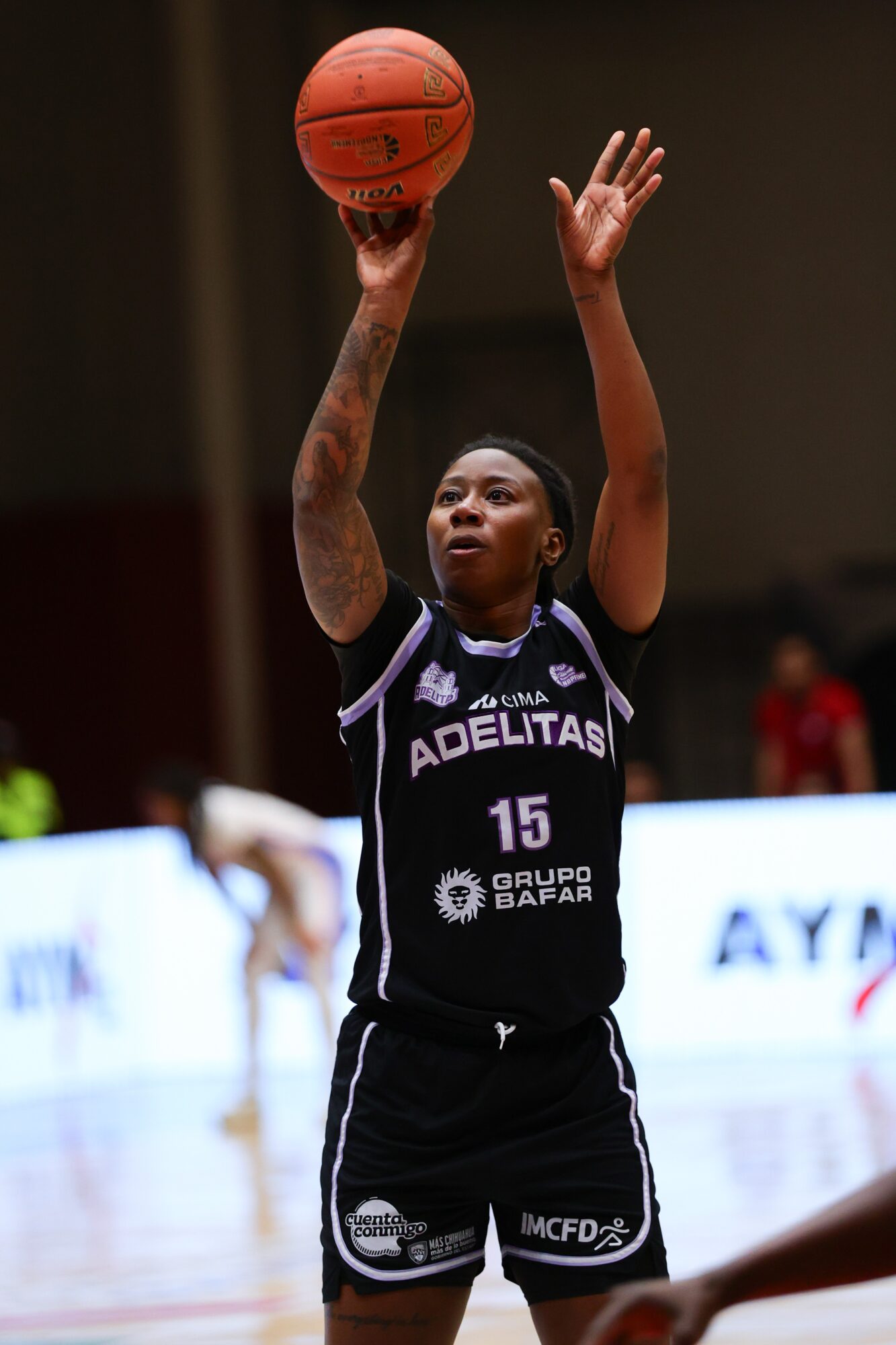 Female basketball player in black uniform shooting a basketball with one arm raised, in an indoor court.