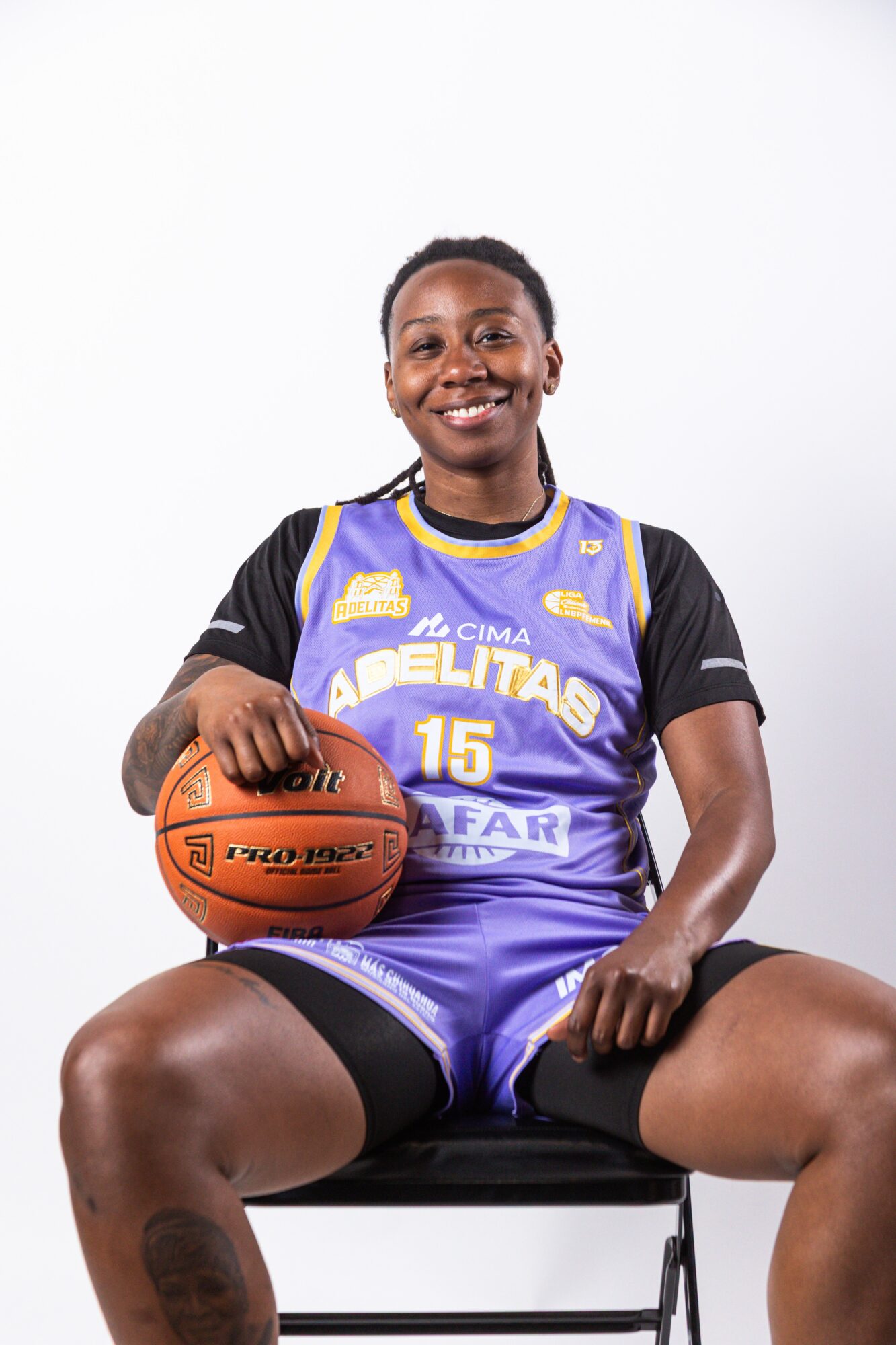 Smiling female basketball player sitting on a chair, holding a basketball, wearing a blue and black uniform, against a white background.
