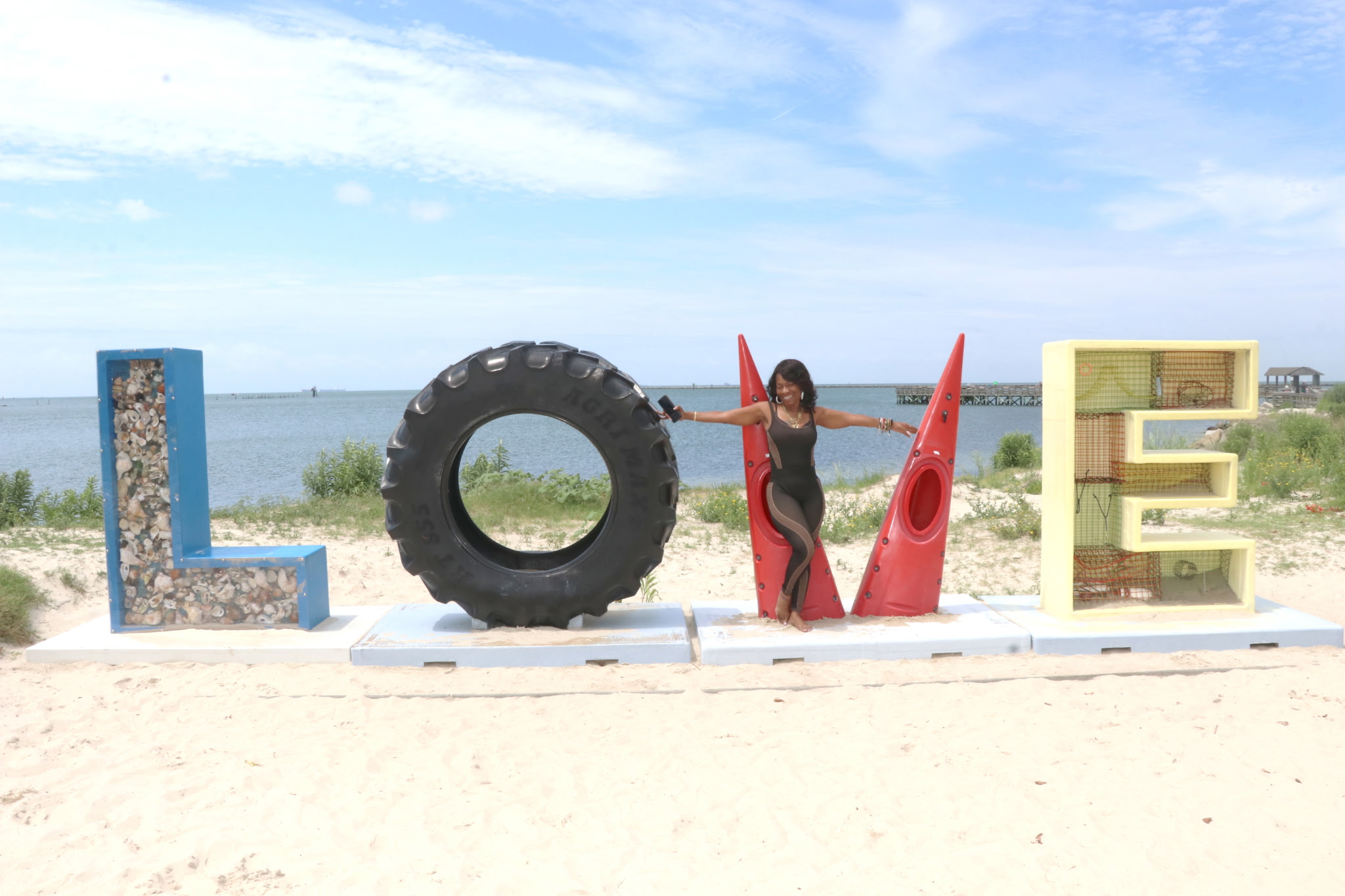 Person standing between large letter sculptures spelling LOVE on a beach, with water and sky in background.