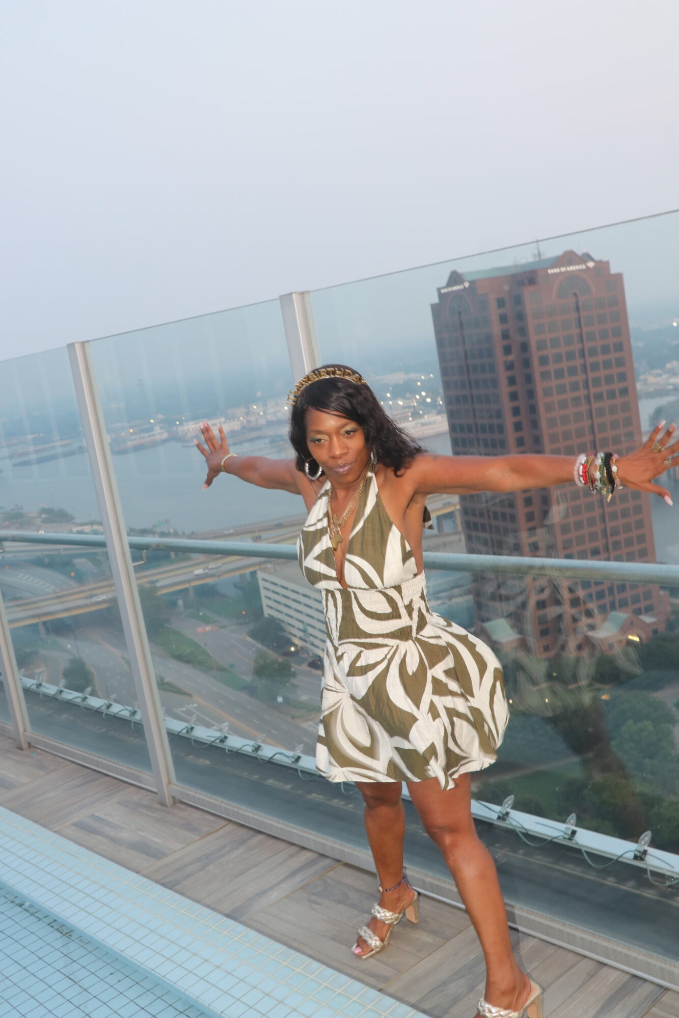 Woman in patterned dress balancing on a balcony with cityscape background, arms outstretched, smiling.