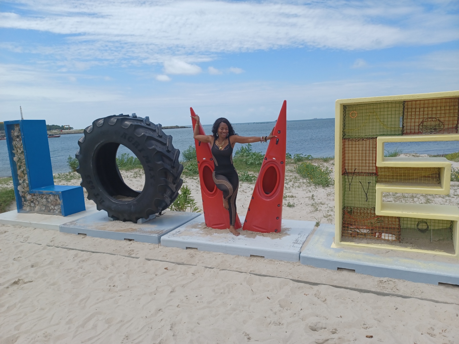 Person sitting between large red sculptures on a beach with water and sky in background.