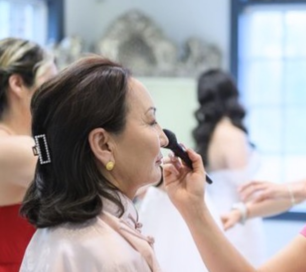 Woman getting makeup applied, with other women in the background, in a bright room.