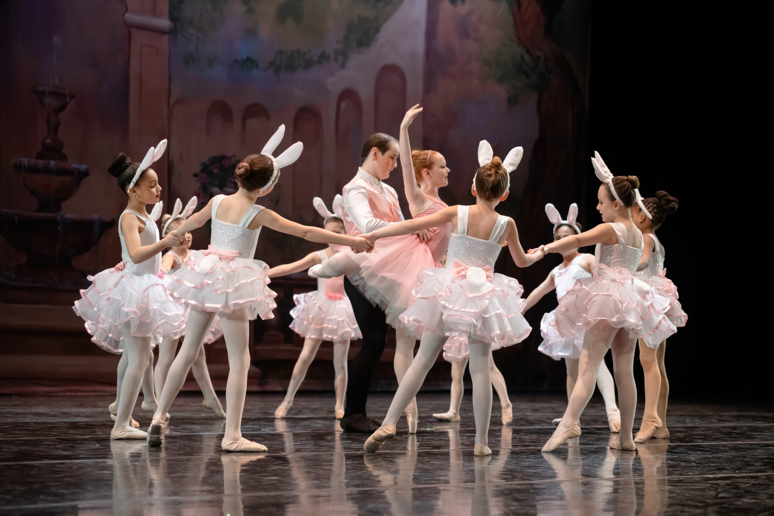 Group of young ballet dancers in pink tutus and bunny ears performing on stage, holding hands in a circle.