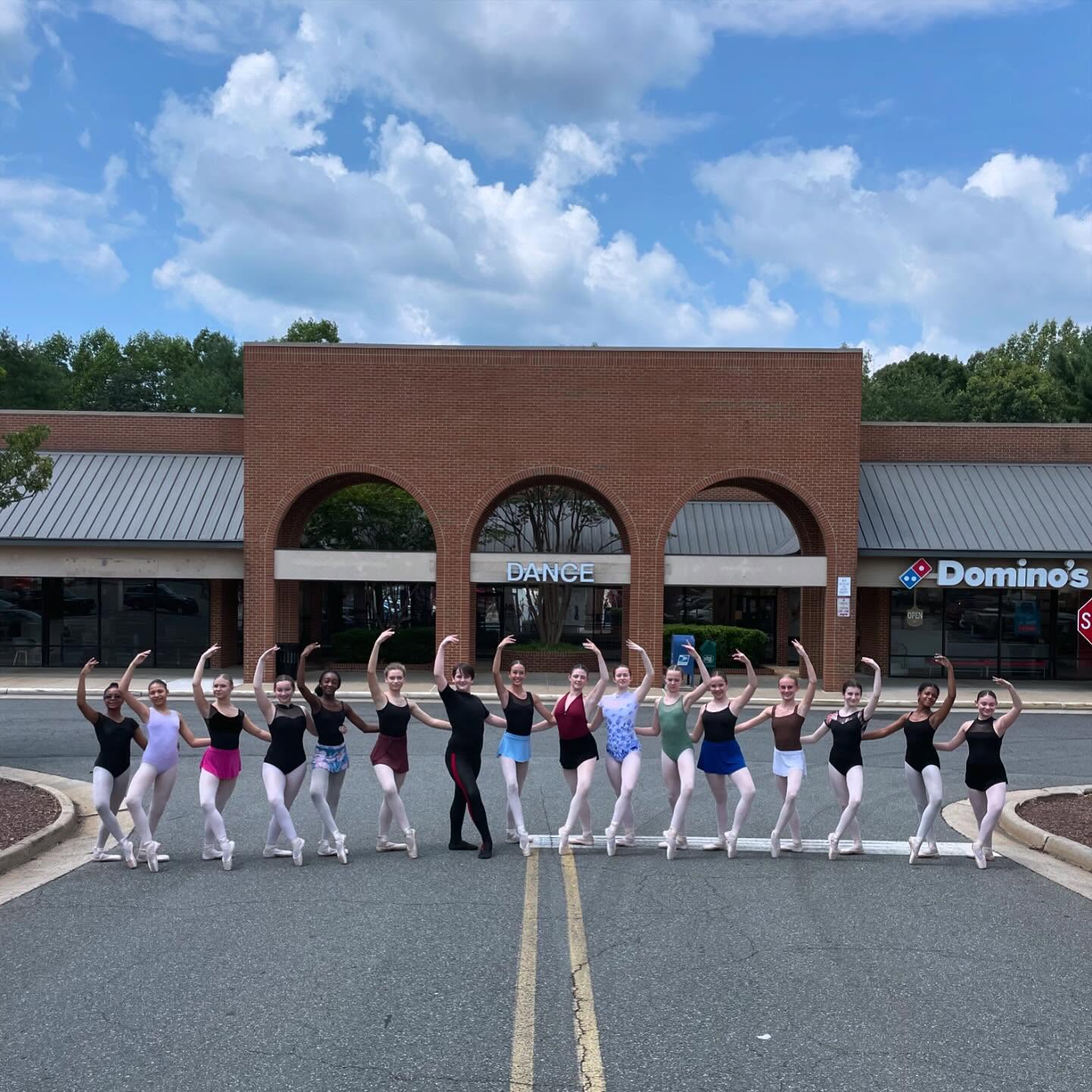 Group of ballet dancers in front of a building with a Domino's Pizza, posing with arms raised, under a partly cloudy sky.