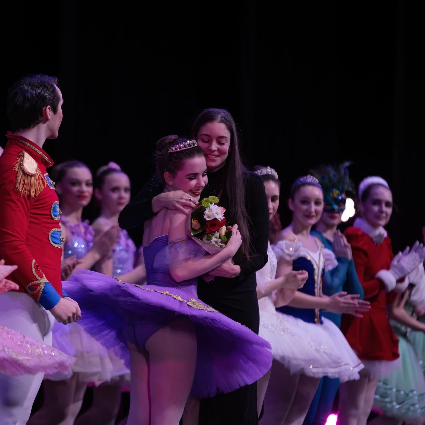 A young girl in a purple ballet costume receives a bouquet from a woman backstage, surrounded by other dancers in costumes.