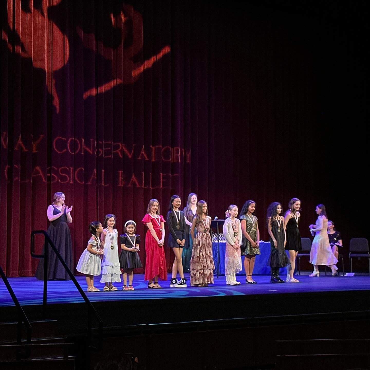 Group of children and adults standing on stage, some holding microphones, in a theater with a red curtain backdrop.