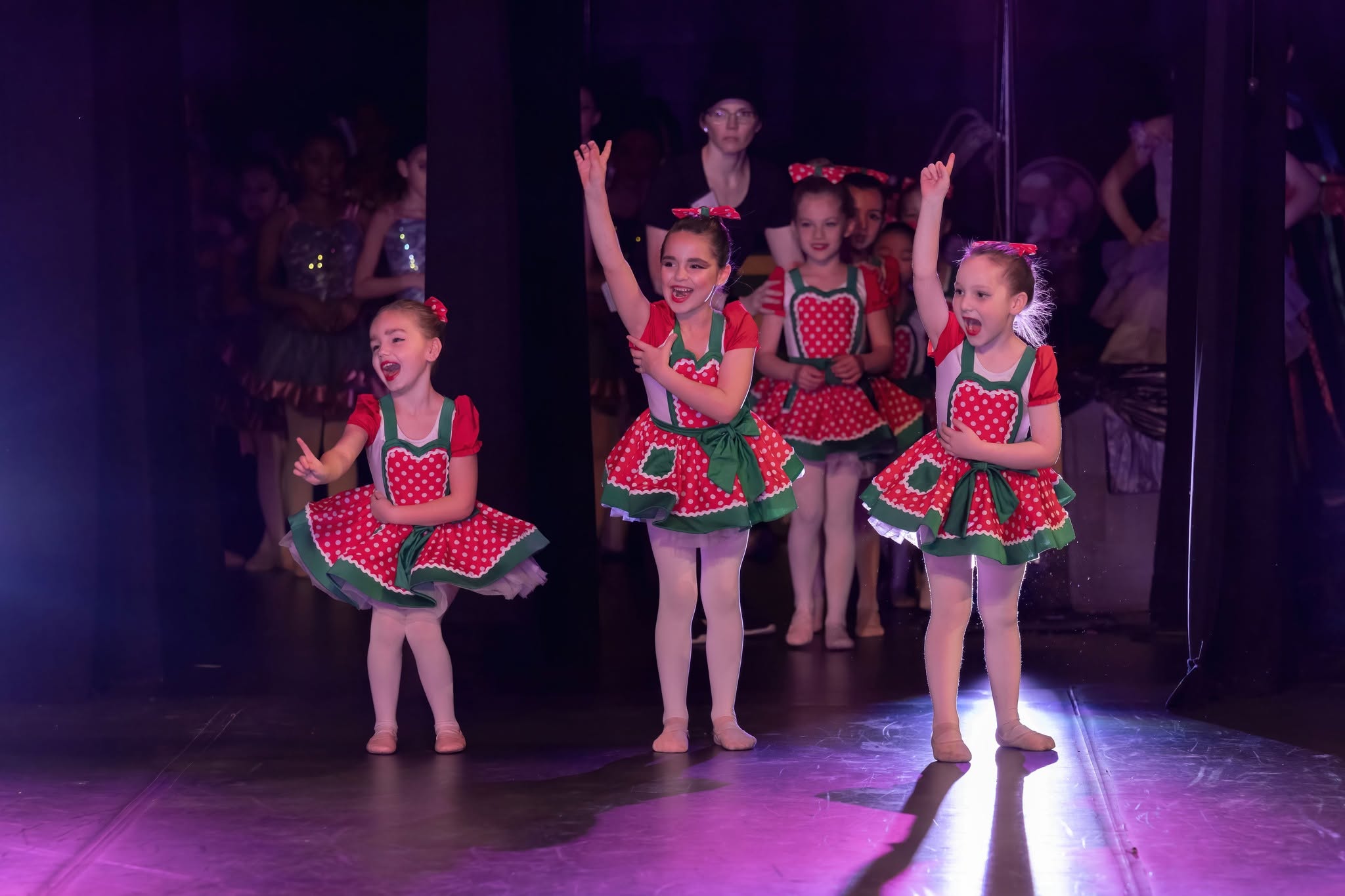 Four young girls in colorful costumes perform on stage, with a dark background and reflections behind them.