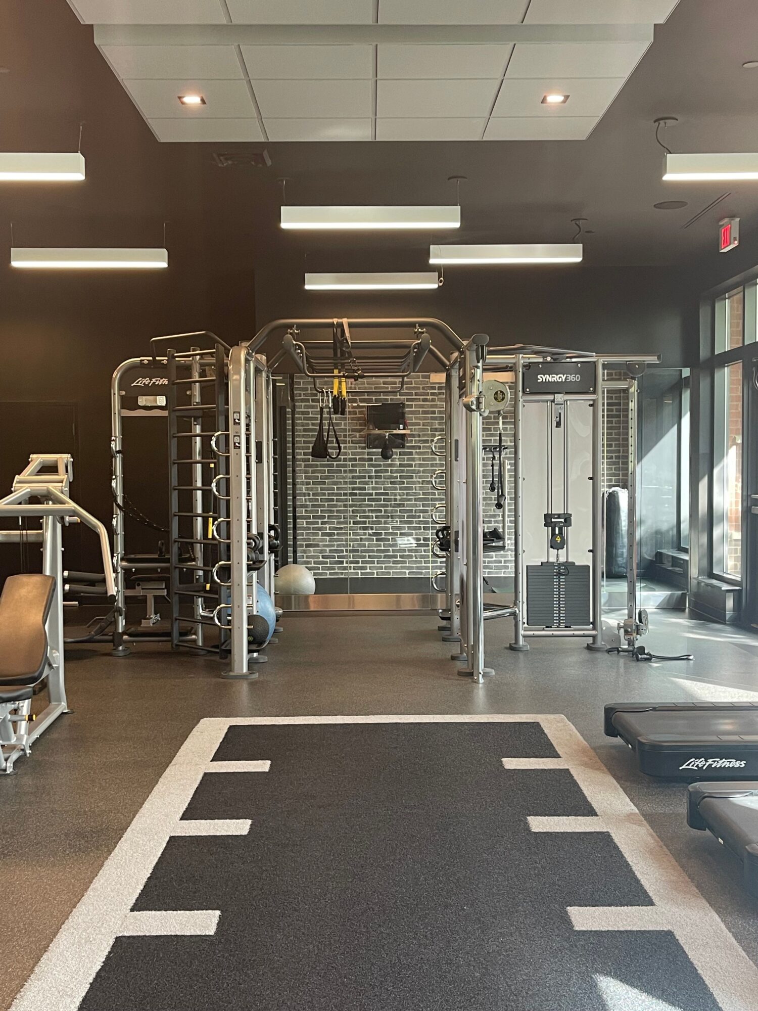 Gym interior with equipment, mirrors, and a black and white patterned floor, viewed from the side. Word 'STRENGTH' visible on the wall.