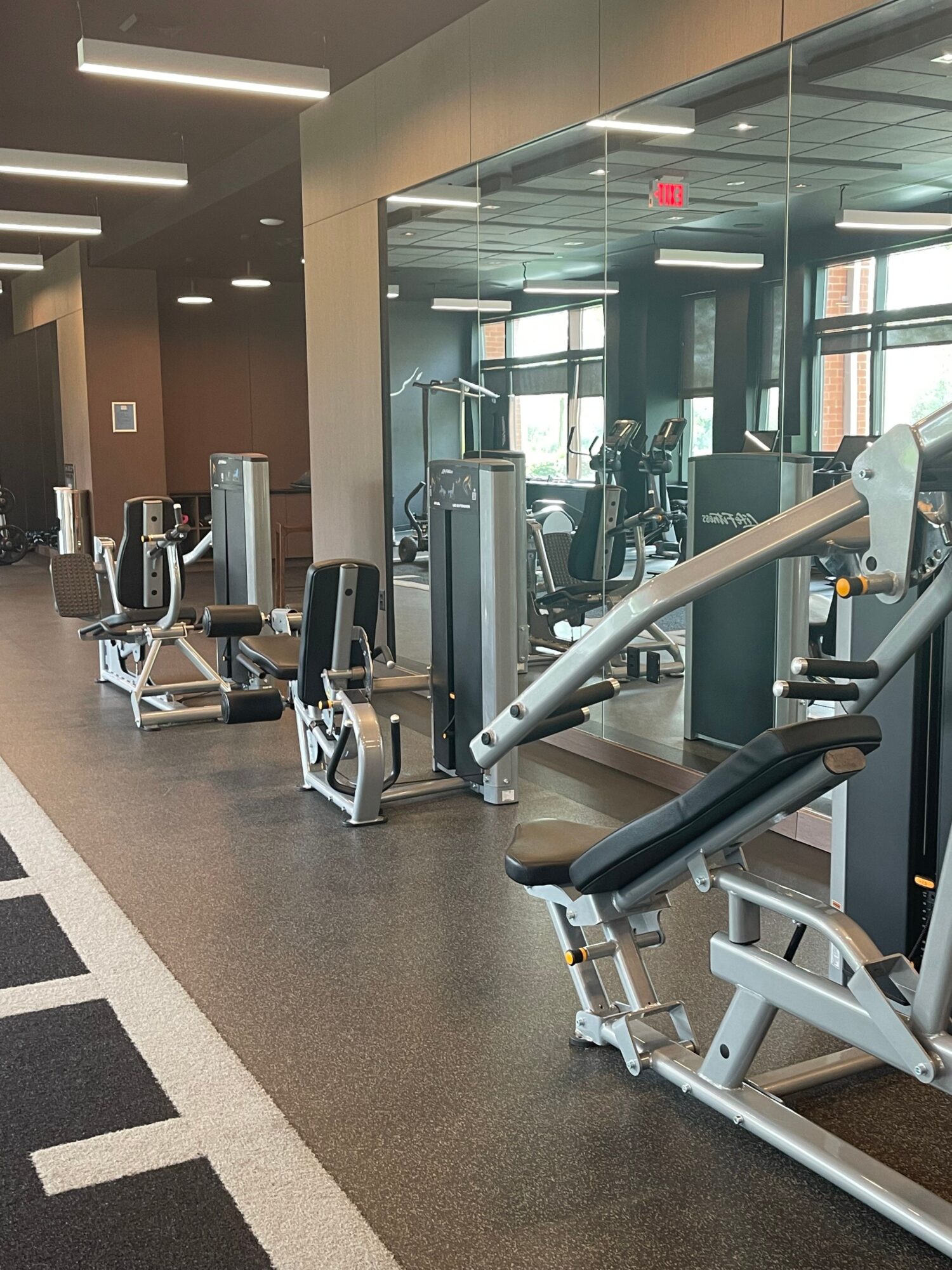 Gym equipment including a seated leg press and other machines in a fitness center with large windows.