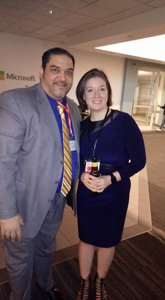 Man and woman standing together indoors, smiling, woman holding a small object, background shows a Microsoft sign.