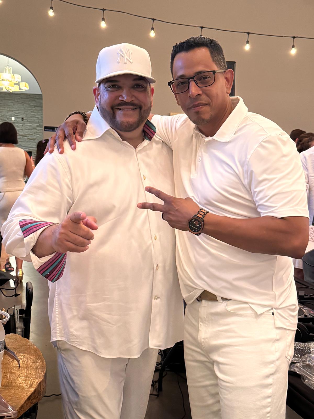 Two men pose together at a social event, one with a white hat and the other with glasses, both in white shirts.