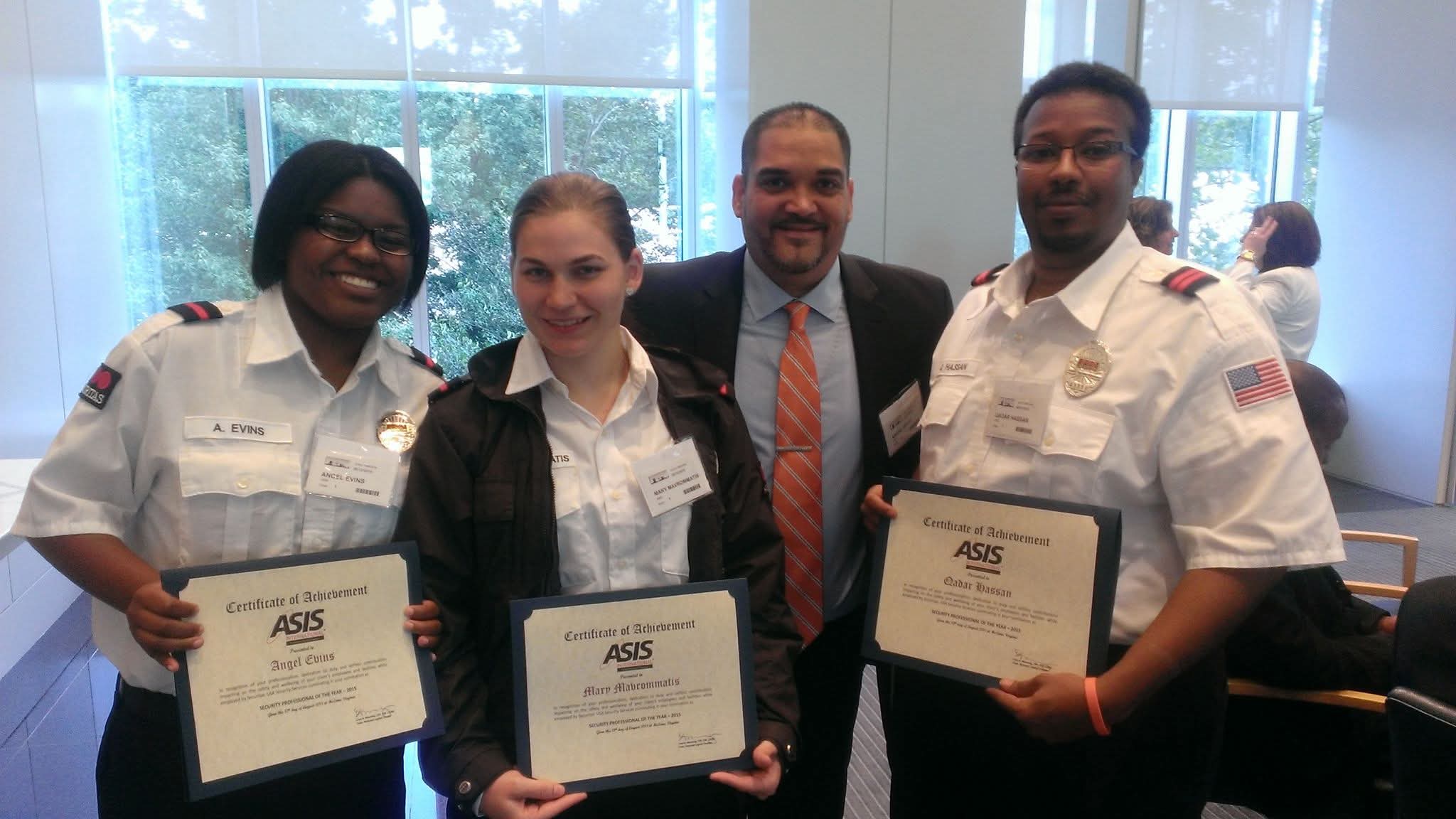 Four people standing indoors, holding certificates, smiling, with windows and other people in the background.