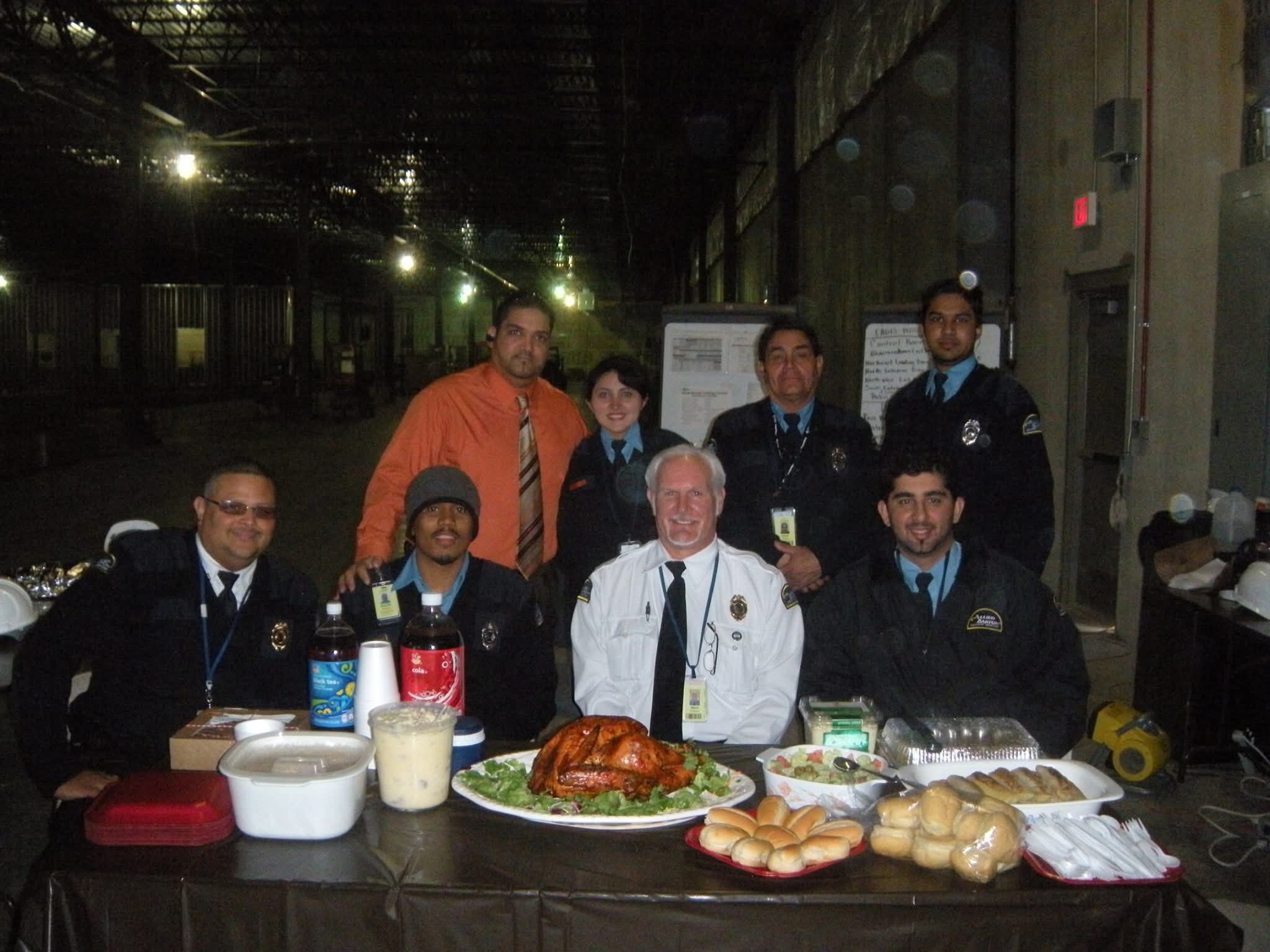 Group of eight people around a table with food, in an industrial setting, some wearing uniforms, one in a white coat.