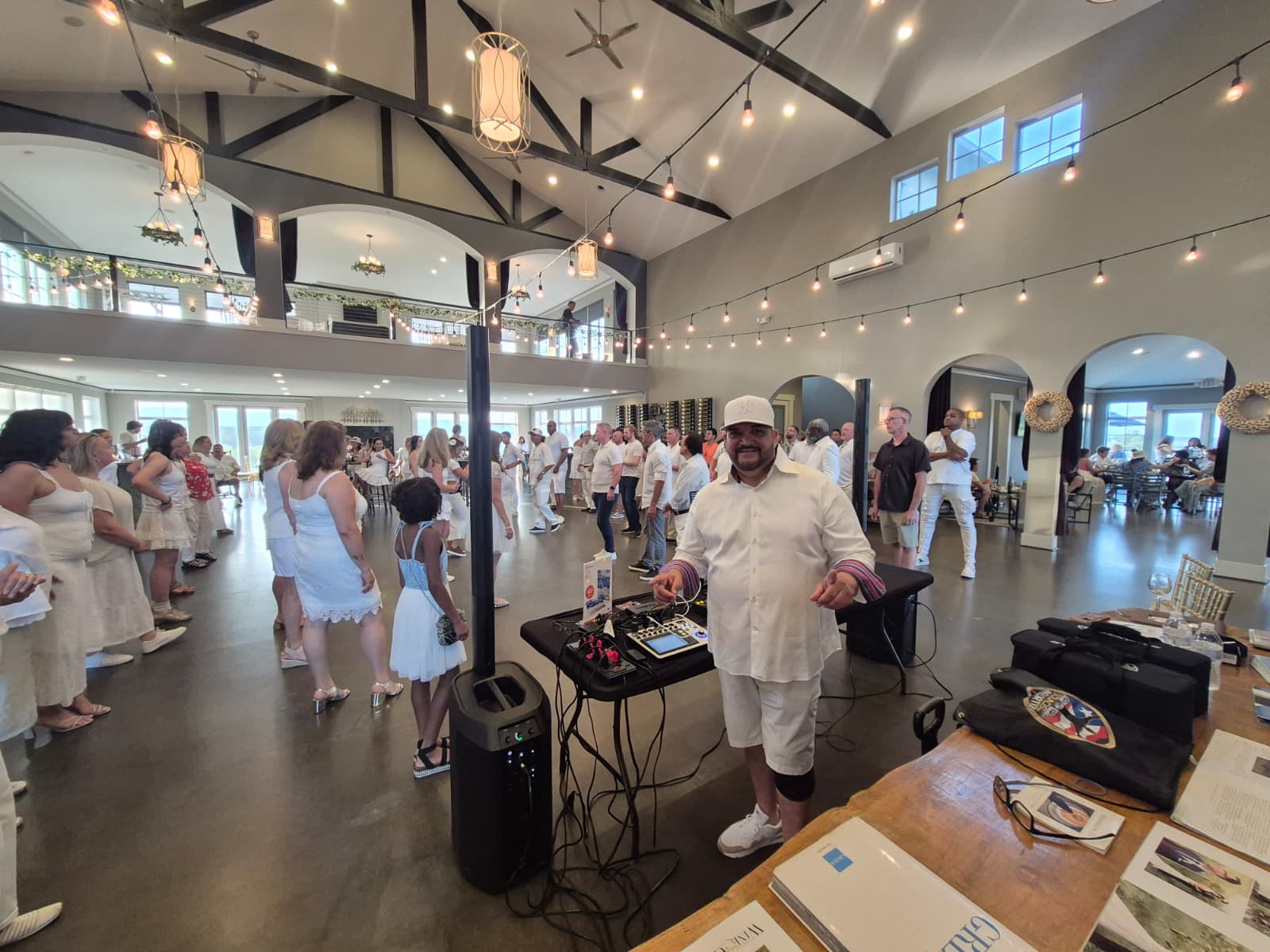 Indoor event with people dancing and socializing, decorated with string lights and hanging lanterns, high ceiling, large windows, and open space.