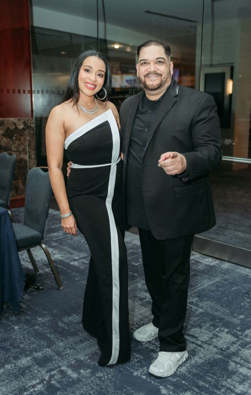 A woman and a man stand together indoors, smiling, with chairs and a glass wall in the background.