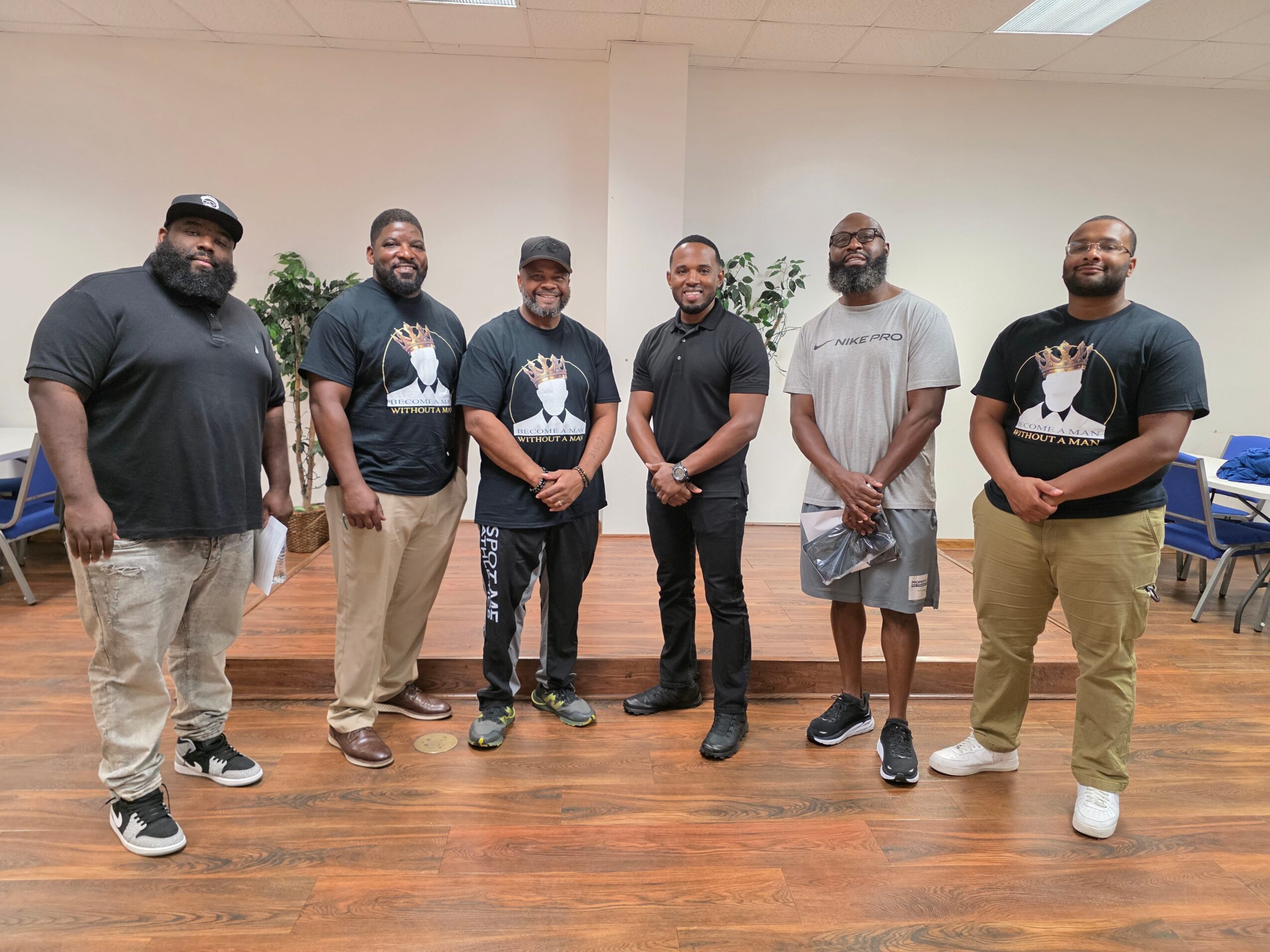Six men standing in a room with wooden floor and white walls, some wearing black T-shirts, some in casual clothing.