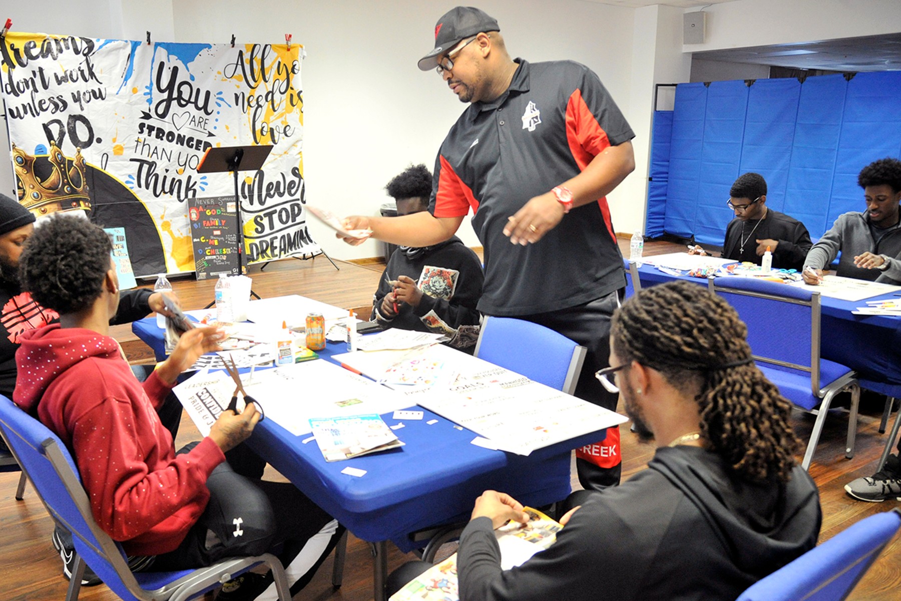 Group of people seated at tables in a classroom, with a man standing and speaking, colorful posters on the wall.
