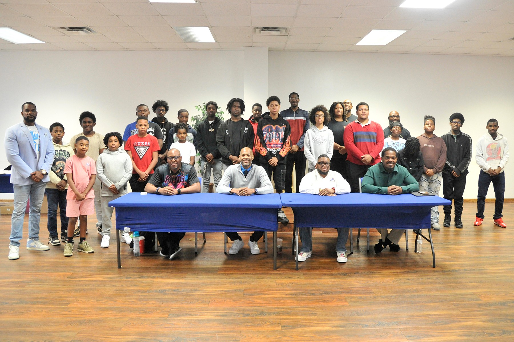 Group of people, including children and adults, gathered in a room with a long table and chairs, some seated and some standing.