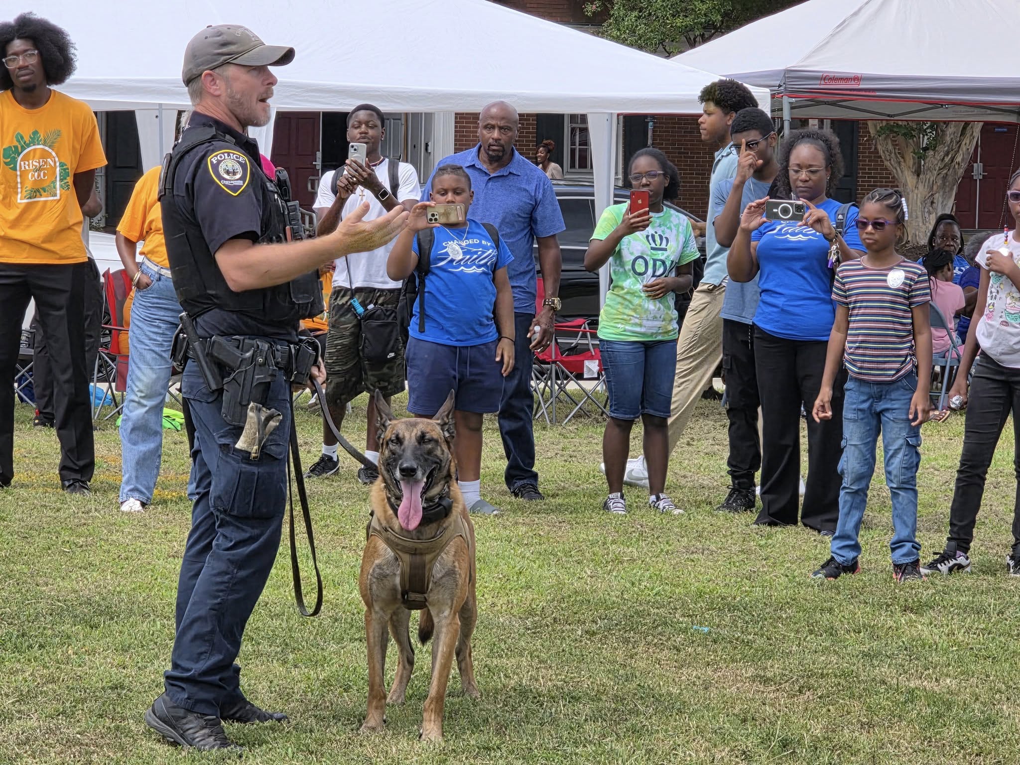 Police officer with a dog in front of a group of people outdoors at an event, tents in background.