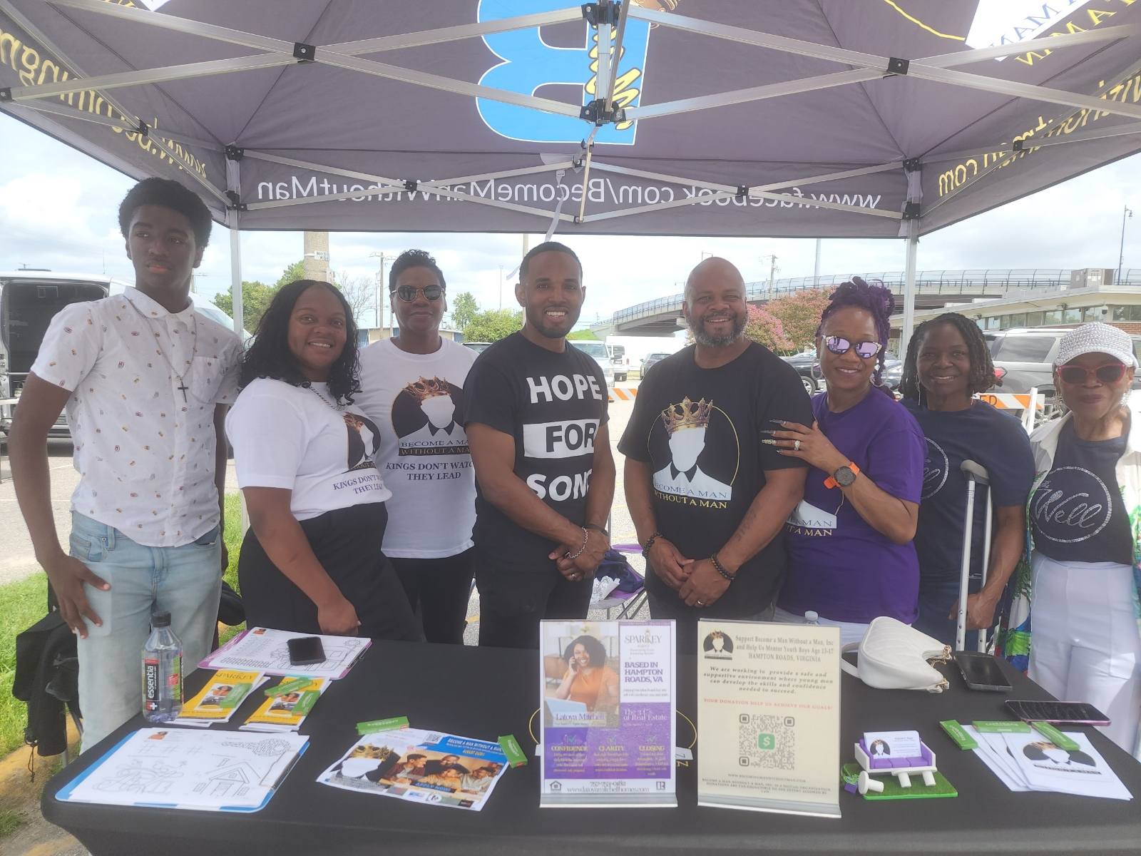 Group of seven people standing under a tent with tables and informational materials, smiling outdoors.