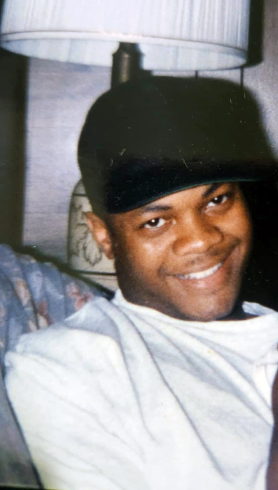 Smiling person wearing a black cap and white shirt, lying on a bed with a lamp above.