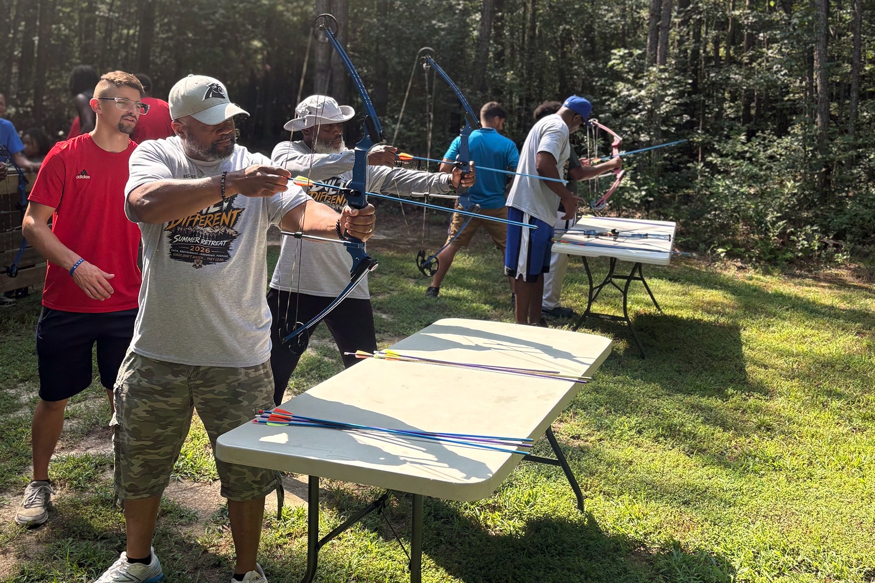 Group of people outdoors participating in archery activities in a wooded area, with tables and equipment visible.