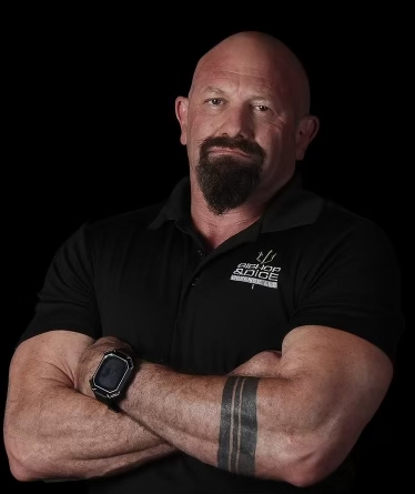 Man with crossed arms, bald head, beard, wearing a black polo shirt and smartwatch, against a black background.